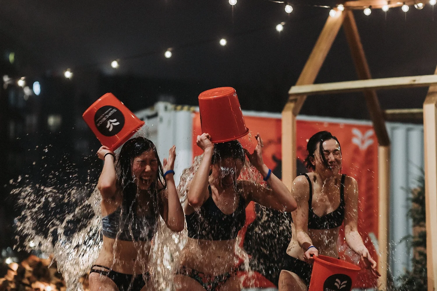 Three women in black swimwear dumping water from red buckets over their heads at night, lit by string lights and a wooden frame in the background.