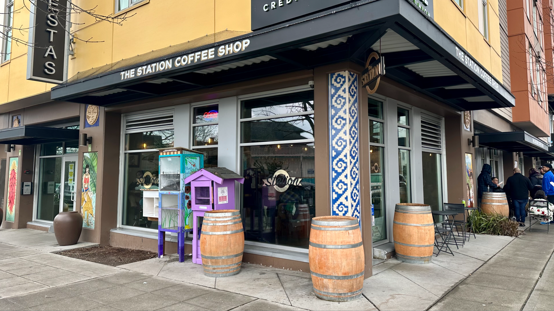 A cornershop coffee house with "The Station Coffee Shop" on a black awning above, and colored tiles on the outer walls.