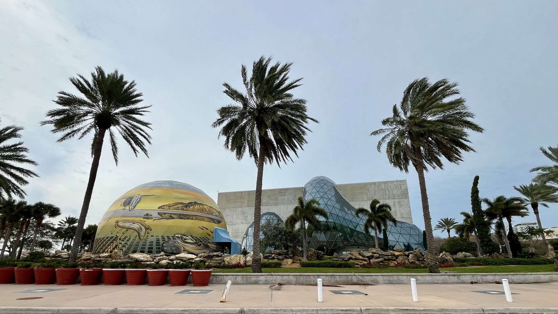 Outdoor scene with palm trees and two distinct buildings: a dome painted with melting clocks artwork and a modern glass structure with a curvy shape under an overcast sky.
