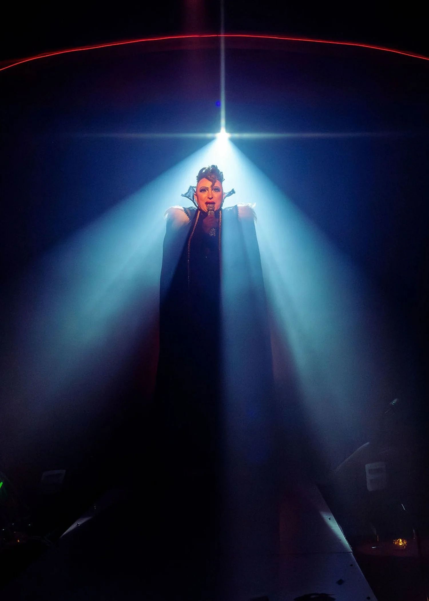 A young man in a dark jacket and red beanie sits at the center of a warmly lit room, holding a microphone as he speaks to an audience. People around him listen attentively, with soft golden light highlighting faces in the crowd.