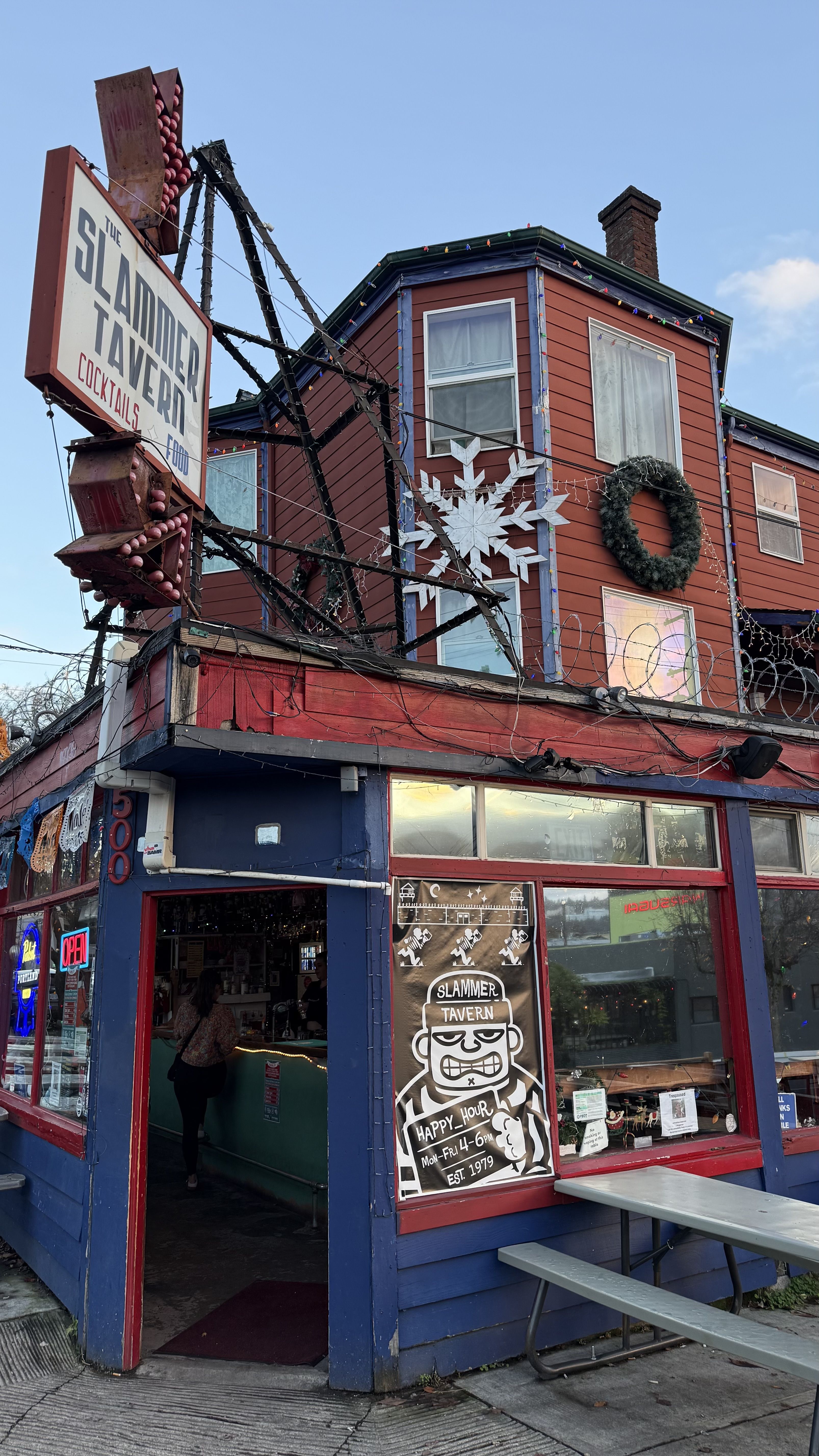 Corner view of a red and blue building with a large sign reading "The Slammer Tavern Cocktails Food." Holiday decorations include a wreath and snowflake on upper windows.
