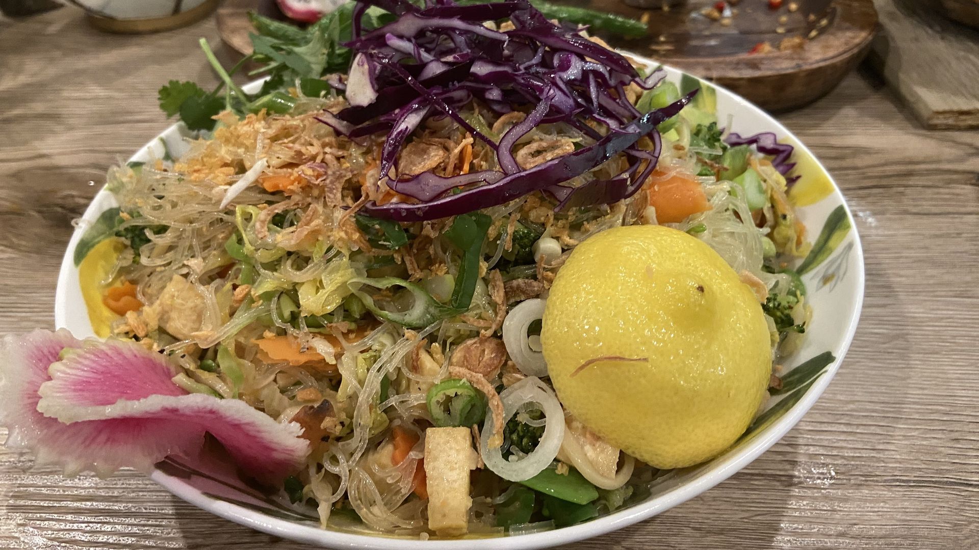 A bowl of glass noodle with shredded purple cabbage, fried onions, and mixed vegetables, plus two lemon halves on a wooden table.