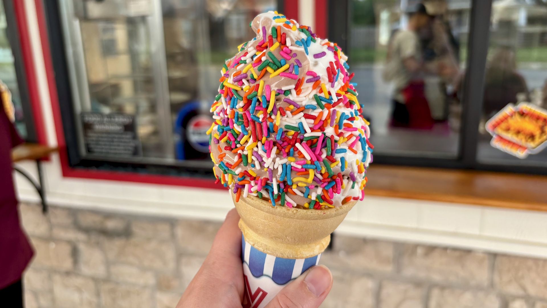 Close-up of an ice cream cone covered in white frosting and colorful rainbow sprinkles, held by a hand; blurred storefront window and people in the background.