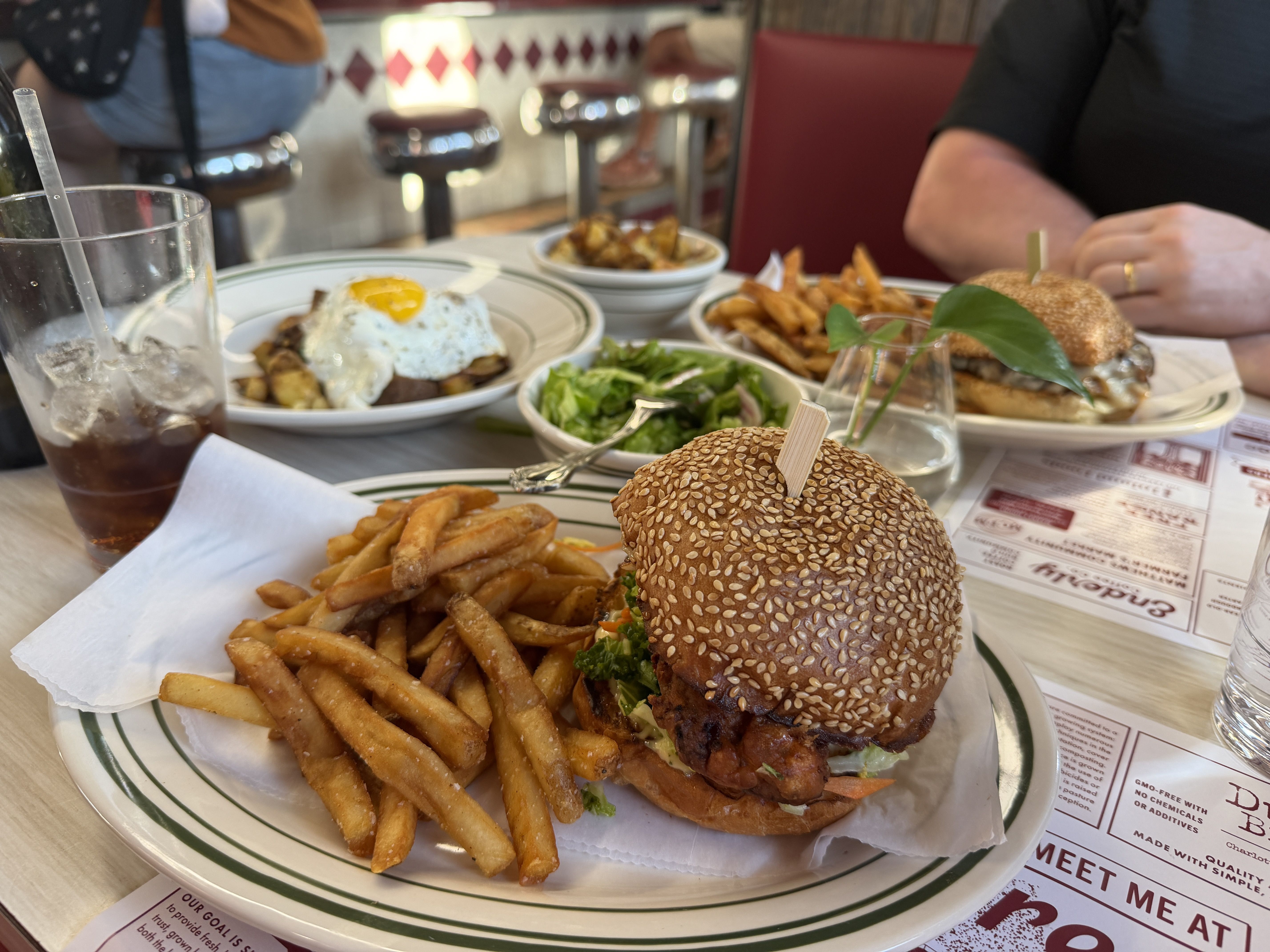 Table scene at a diner: two sesame-seed burgers with fries, a side salad, and a glass of iced cola, with a person in a black shirt in the background amid red booth seating.