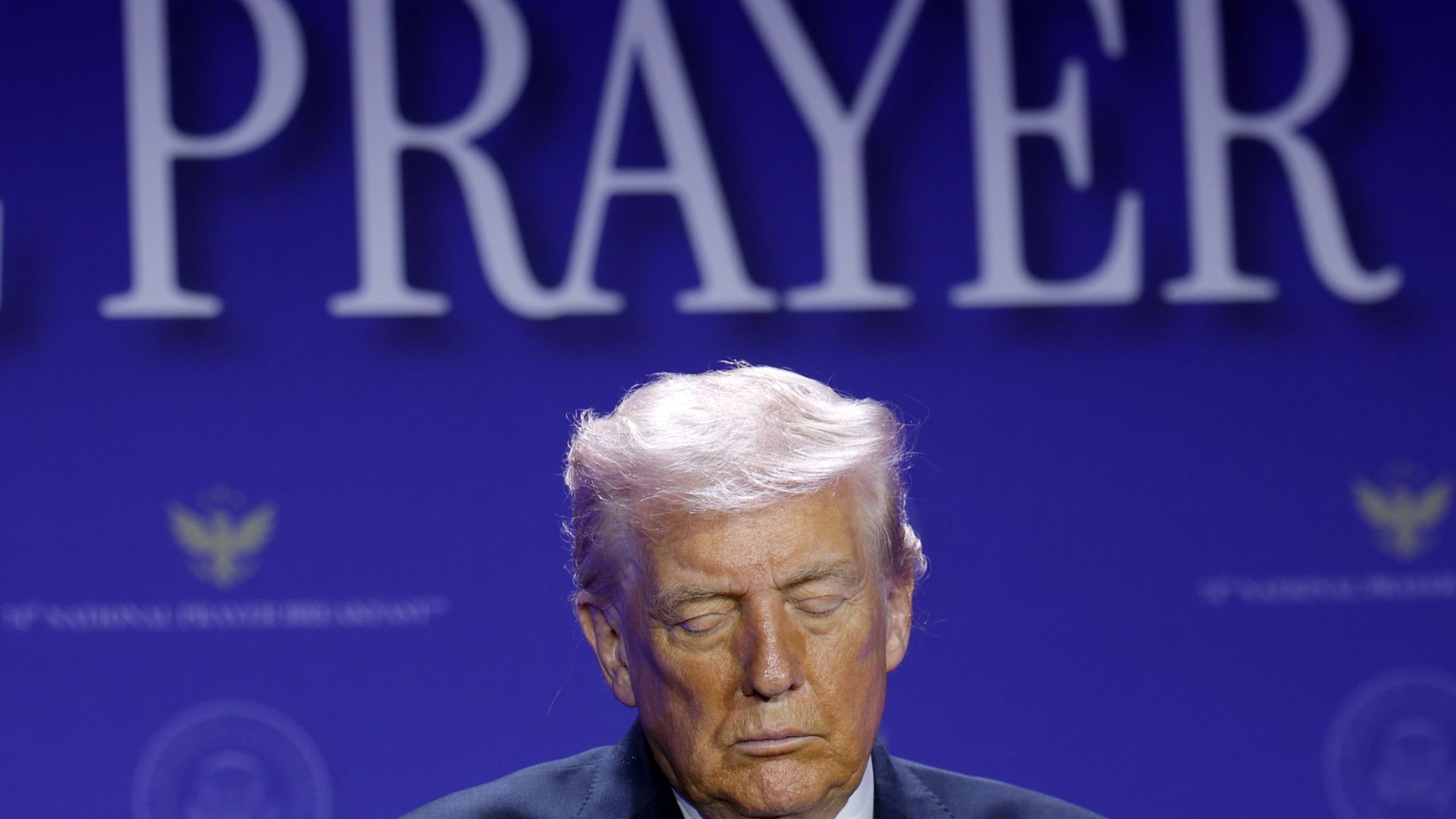 President Trump in a dark suit stands on a stage, eyes closed, against a blue backdrop with large white letters reading "PRAYER" behind him.