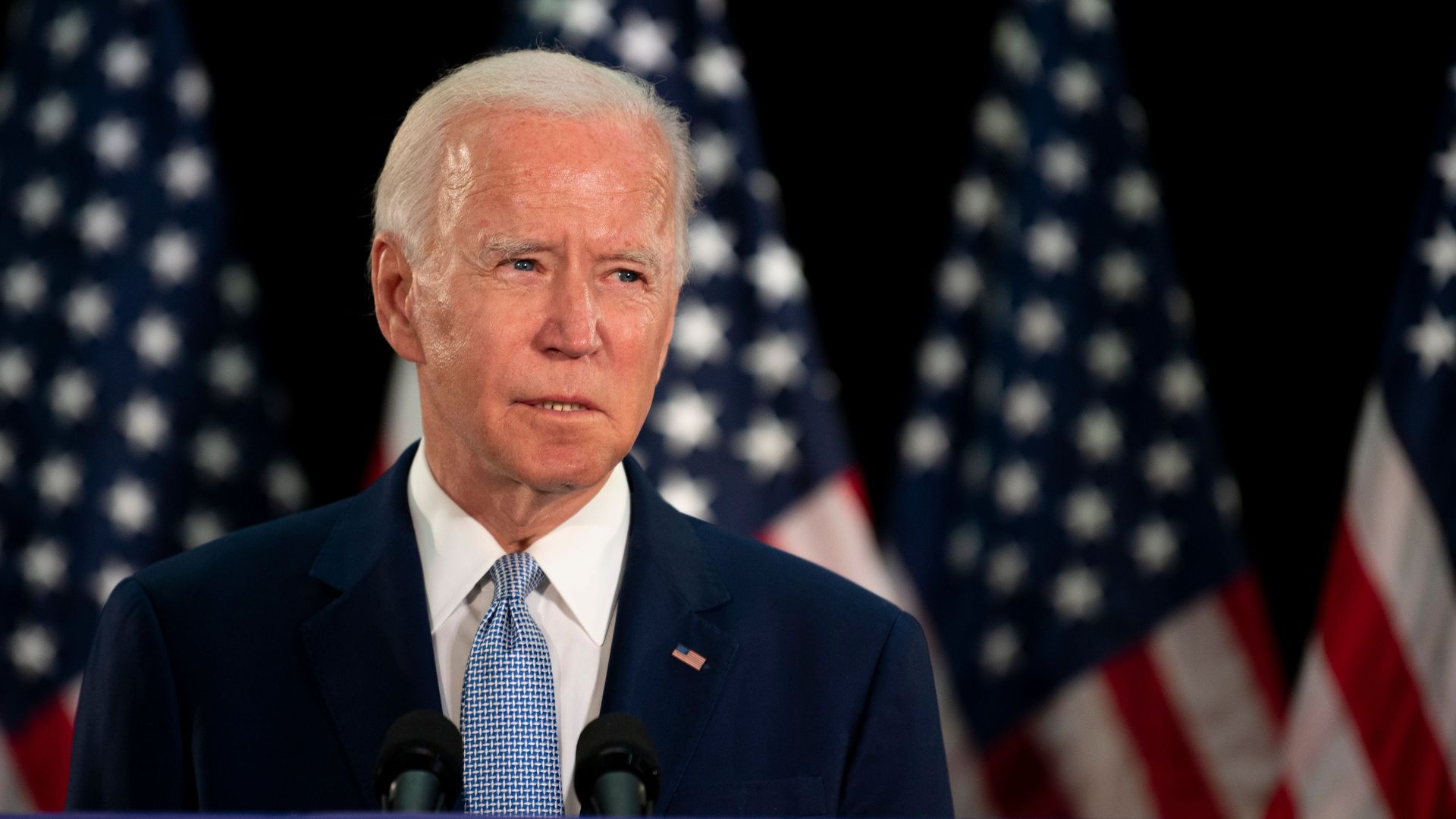 Joe Biden stands in front of American flags in a suit