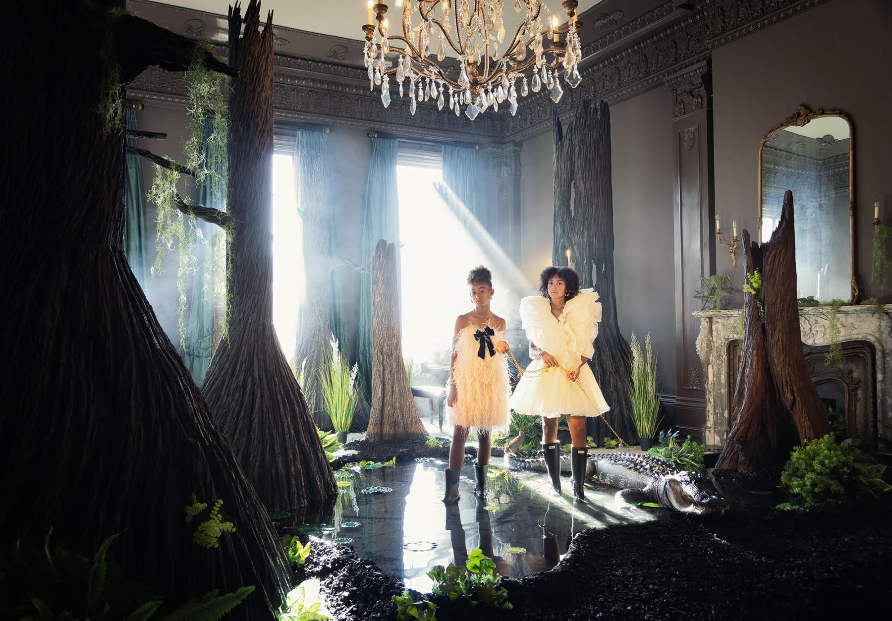 Two girls in white dresses and black boots stand on water surrounded by large tree trunks and plants in an ornate room with sunlight streaming through windows.