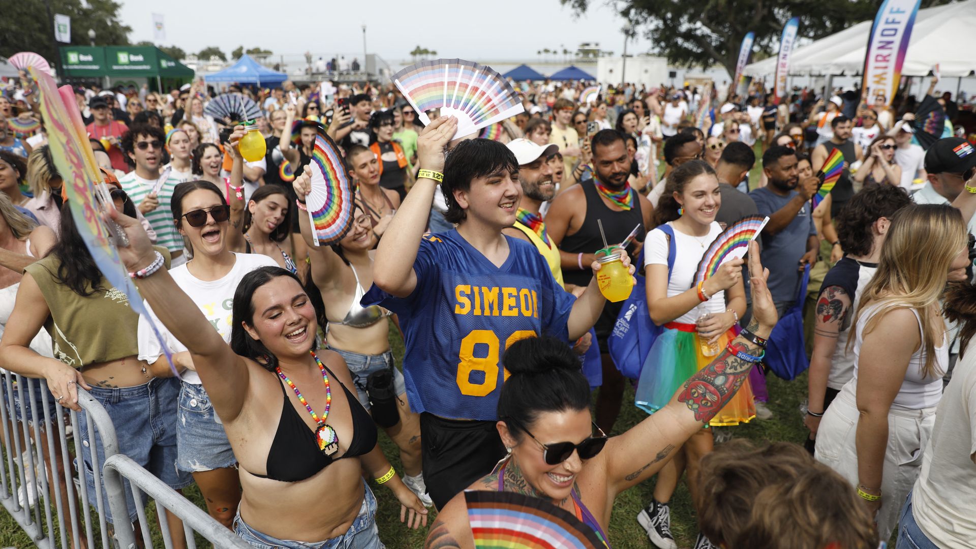 Vibrant Pride festival crowd outdoors: people waving rainbow fans, wearing colorful outfits and beads. A man in a blue jersey holds a large rainbow fan at center, tents in background.