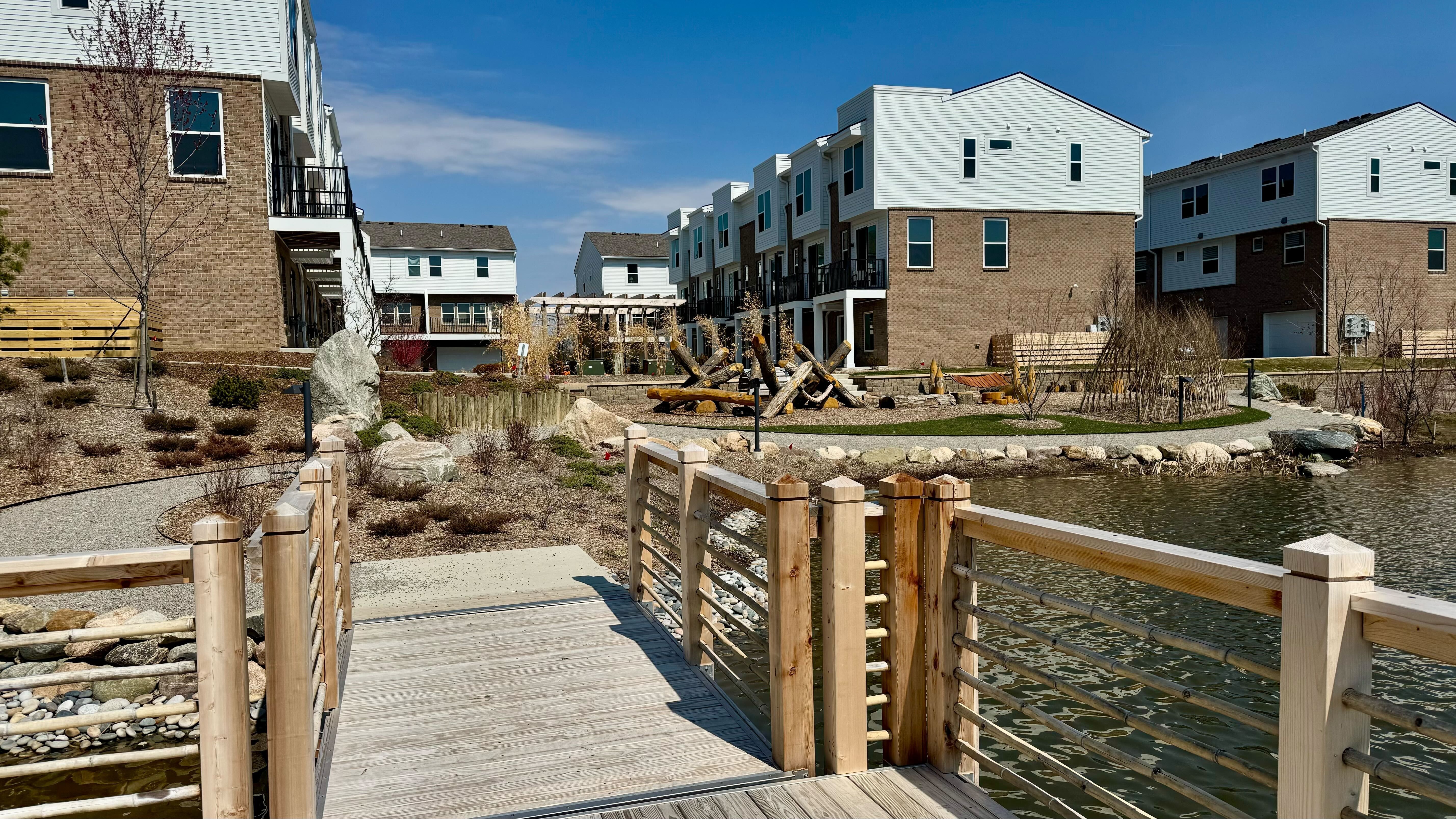 Sunlit residential courtyard with brick-and-white townhomes, balconies, and a wooden boardwalk over a pond. Rocky landscaping and leafless trees frame a central pile of logs 