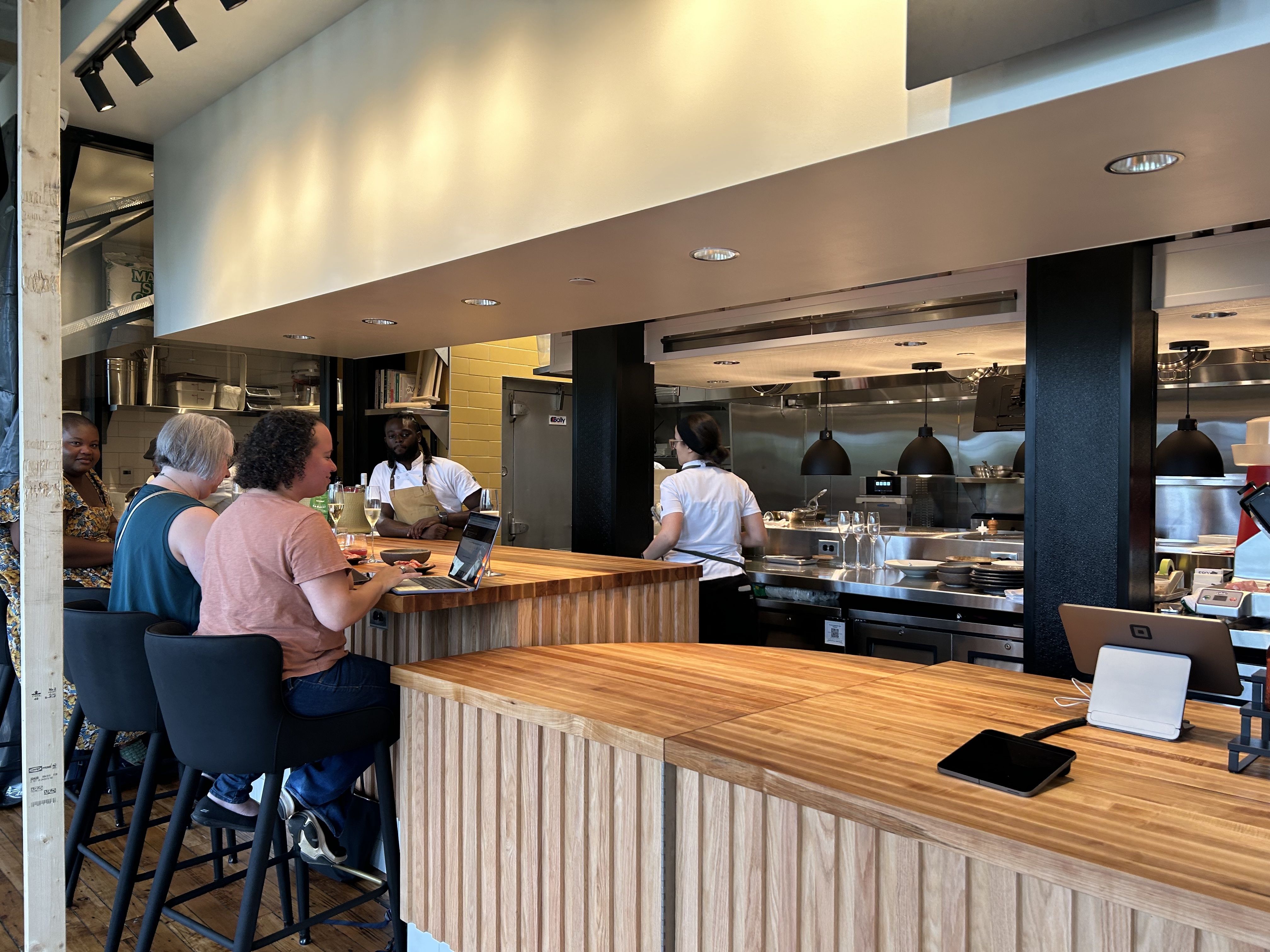 People sitting at a wooden bar counter in a modern kitchen area, with chefs working behind the counter and a laptop and drinks on the bar.