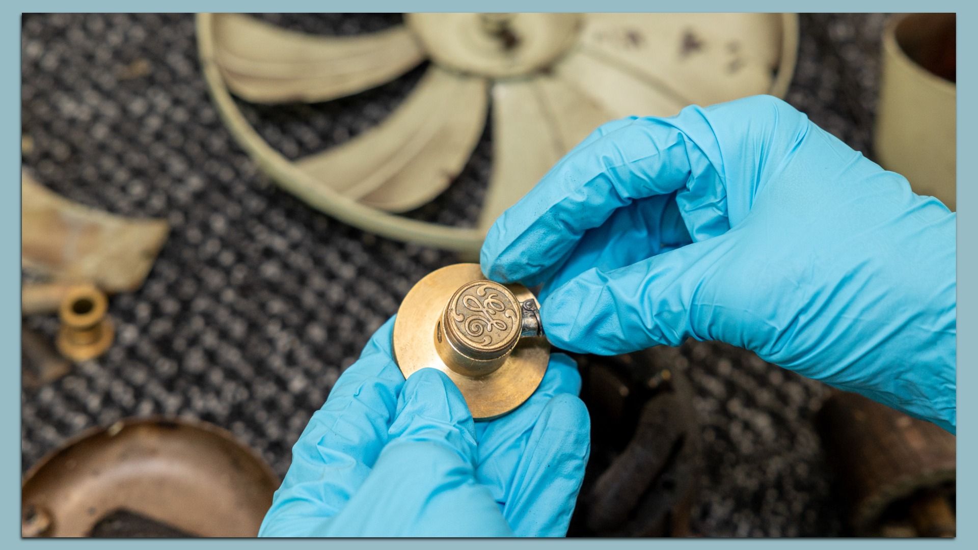 Close-up of blue-gloved hands holding a brass circular emblem with an ornate engraving; blurred workshop machinery in the background.