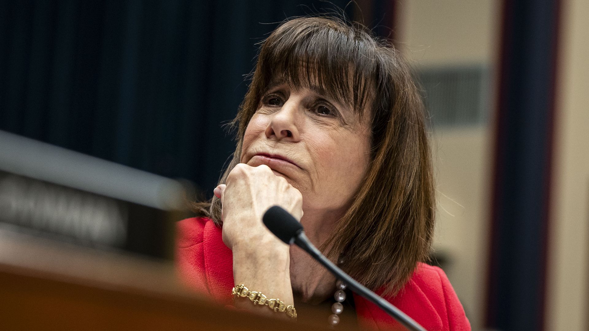 Rep. Kathy Manning, wearing a red blazer, bracelet and necklace, leaning her chin on her fist while sitting at a committee dais microphone.