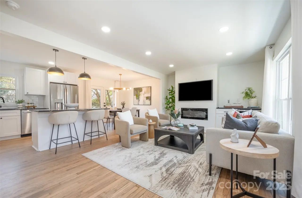 Bright open living room and kitchen with white walls, wooden floors, beige armchairs, a gray couch with pillows, black coffee table, modern pendant lights, and stainless steel appliances.
