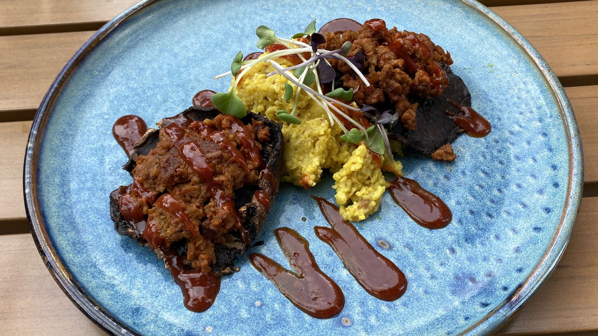 A blue plate with cheesy grits, two mushrooms topped with imitation meat and drizzled barbecue sauce