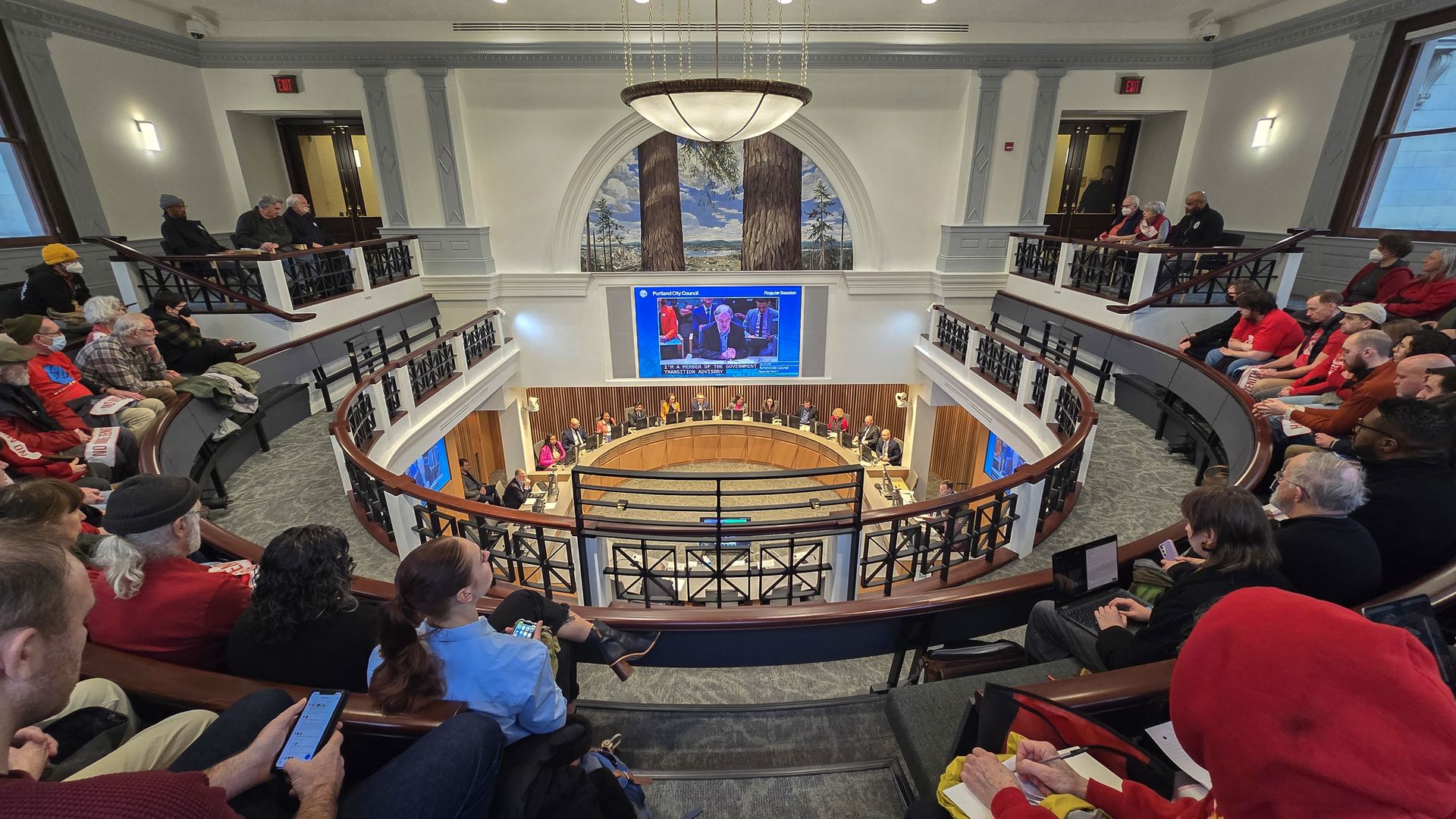 People seated in a circular council chamber watching a meeting with officials at a round wooden desk, screens showing speakers, and a mural of tall trees on the wall.
