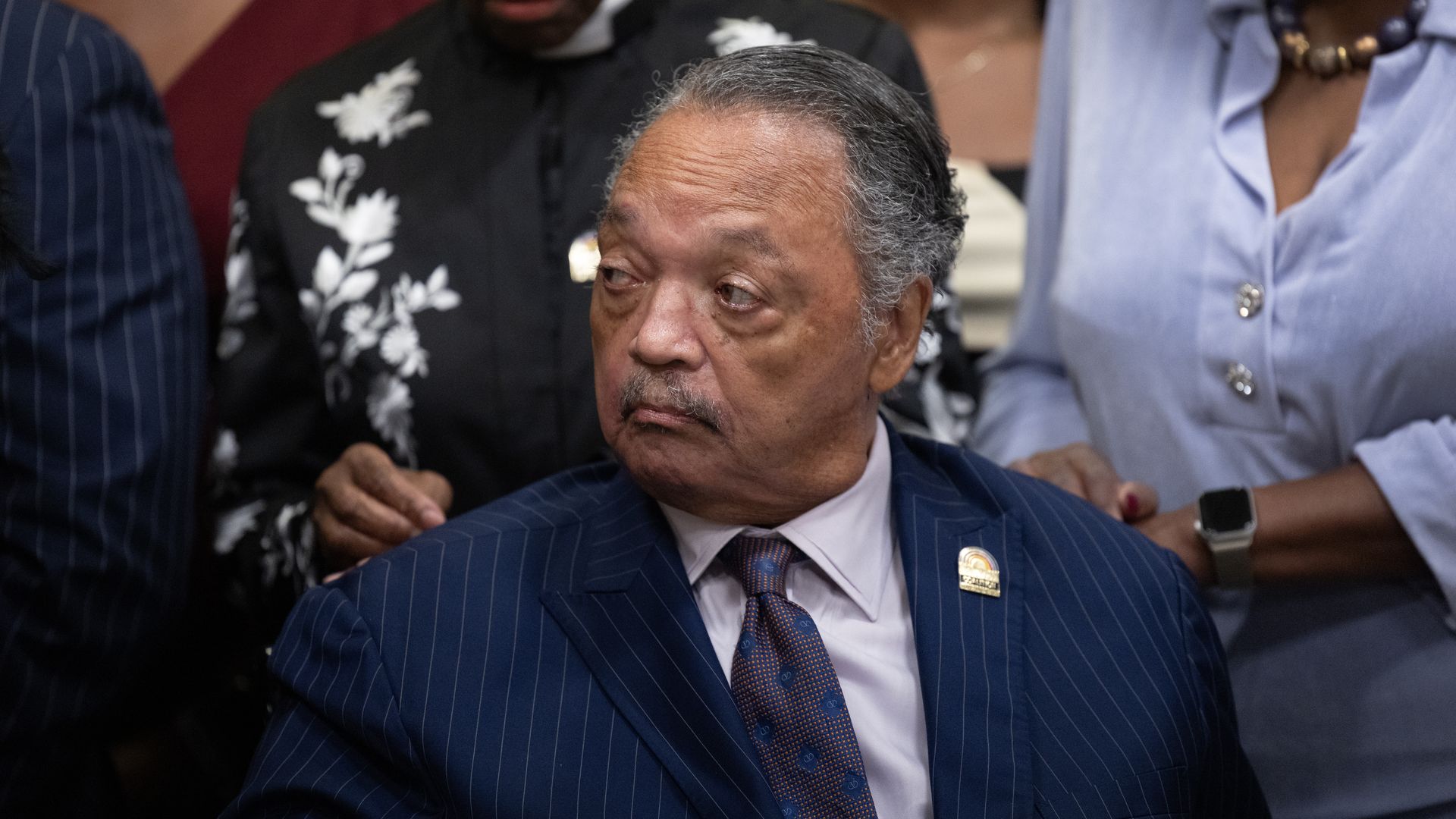 Older man in a blue pinstripe suit and patterned tie looks to the side, surrounded by people wearing black, blue, and maroon clothing in a close indoor setting.