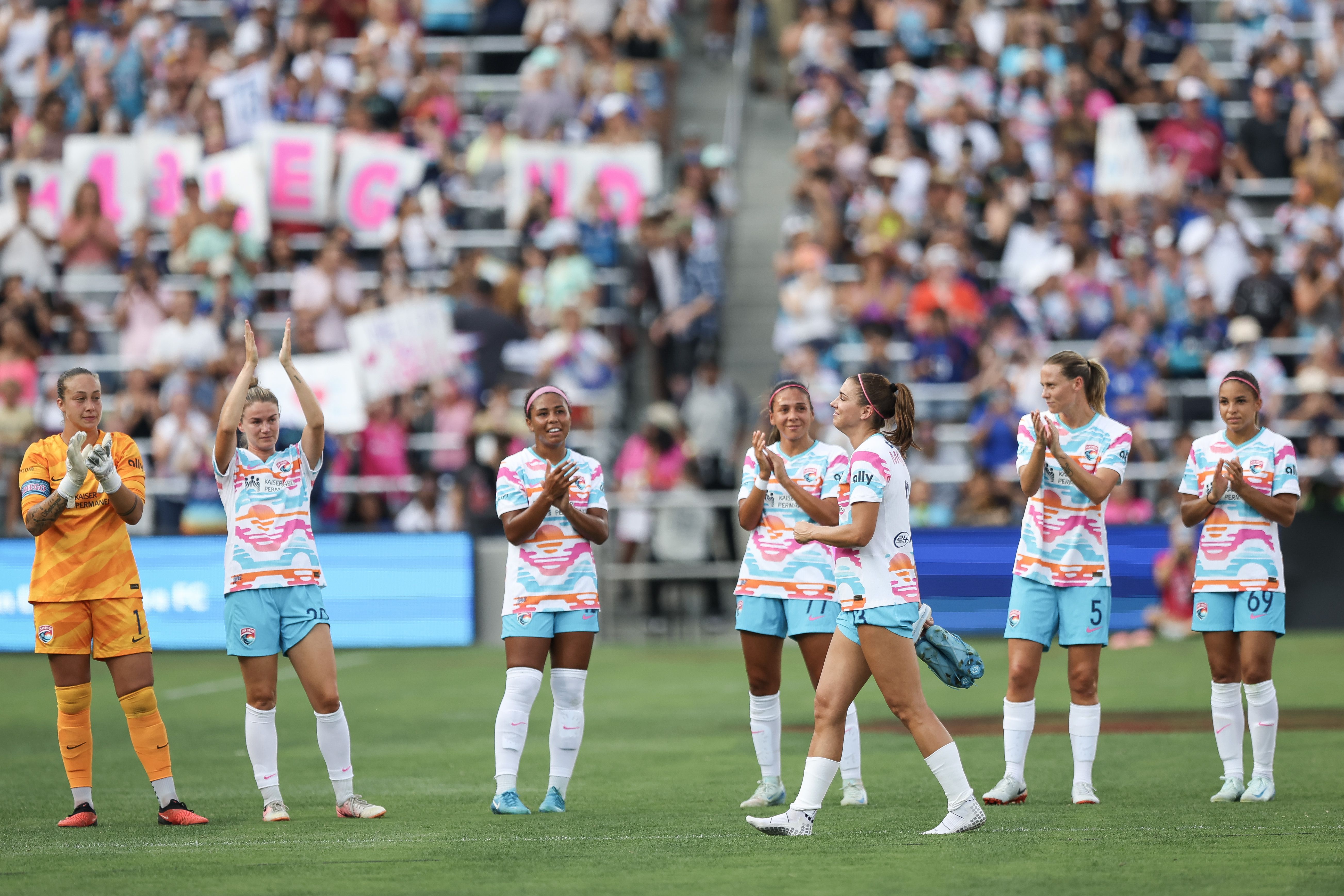 A woman takes off her cleats and walks off a soccer field with people clapping in the background