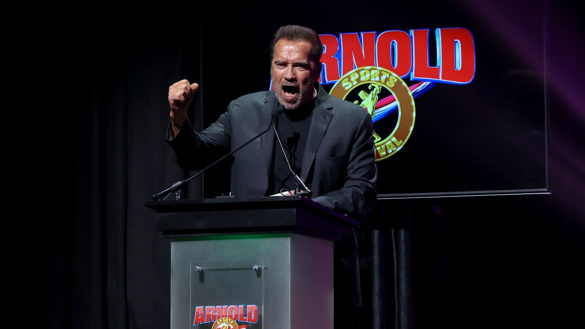 Arnold Schwarzenegger holds up a fist while speaking at a podium