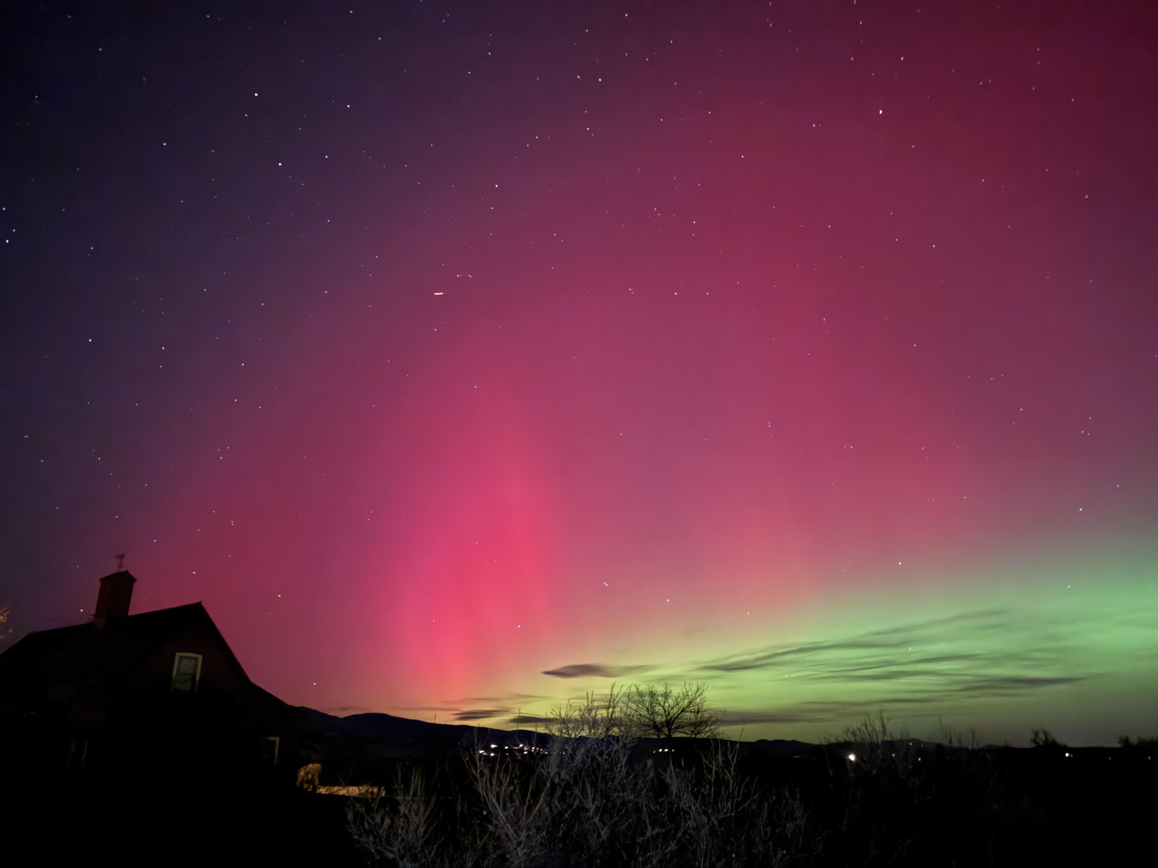 Night sky with bright red and green aurora borealis above dark silhouette of a house and trees, scattered stars visible in the purple gradient sky.