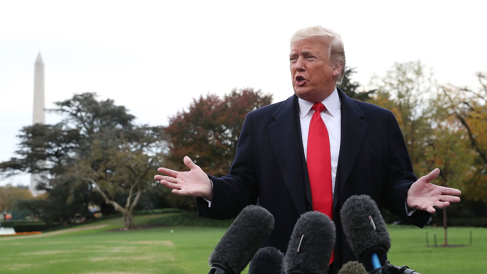Donald Trump hands out in shrug with mics before him and Washington Monument behind.