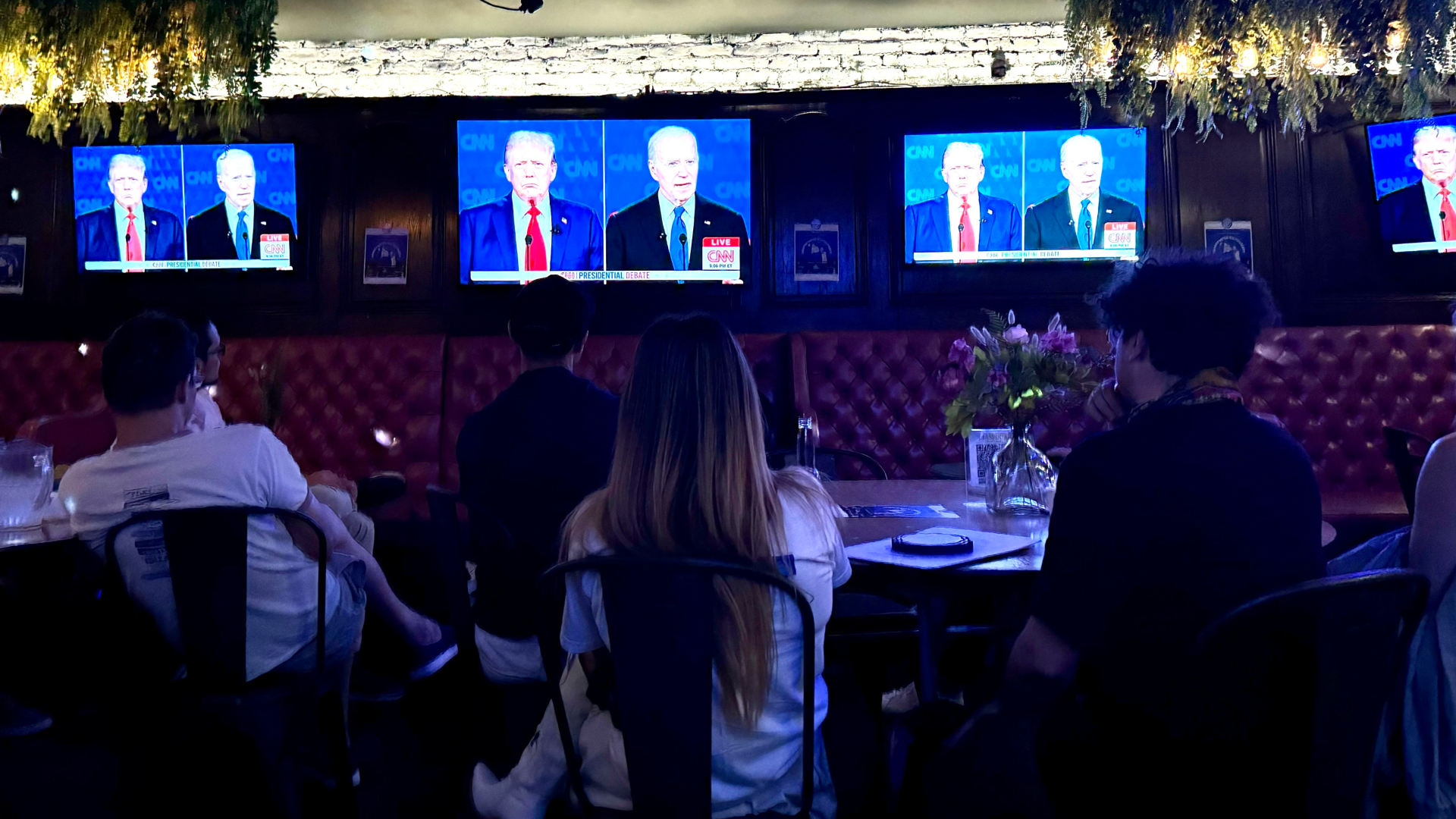 People sitting in chairs watching a set of TVs playing the presidential debate with a side-by-side of former president Donald Trump and President Joe Biden. 