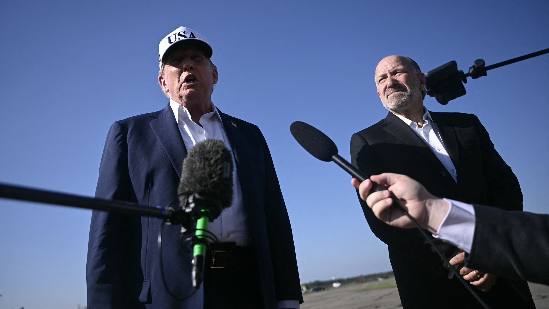 President Trump and Commerce Secretary Howard Lutnick in suits stand outdoors under a clear blue sky, one wearing a white "USA" cap speaking into microphones held by unseen hands.