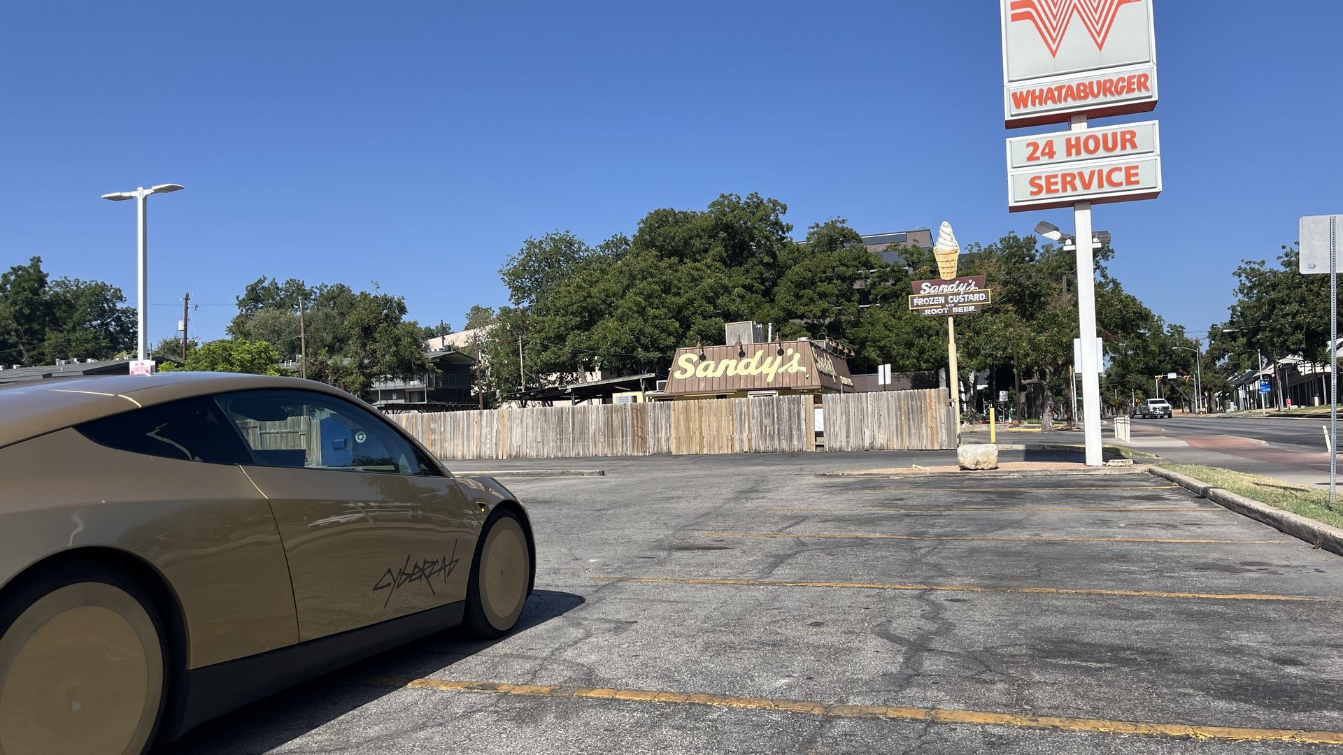 Clear blue sky over a parking lot. A tan car sits in the left foreground. A tall Whataburger sign with an orange W and the words "24 HOUR SERVICE" rises on the right, with trees and Sandy's in the background.