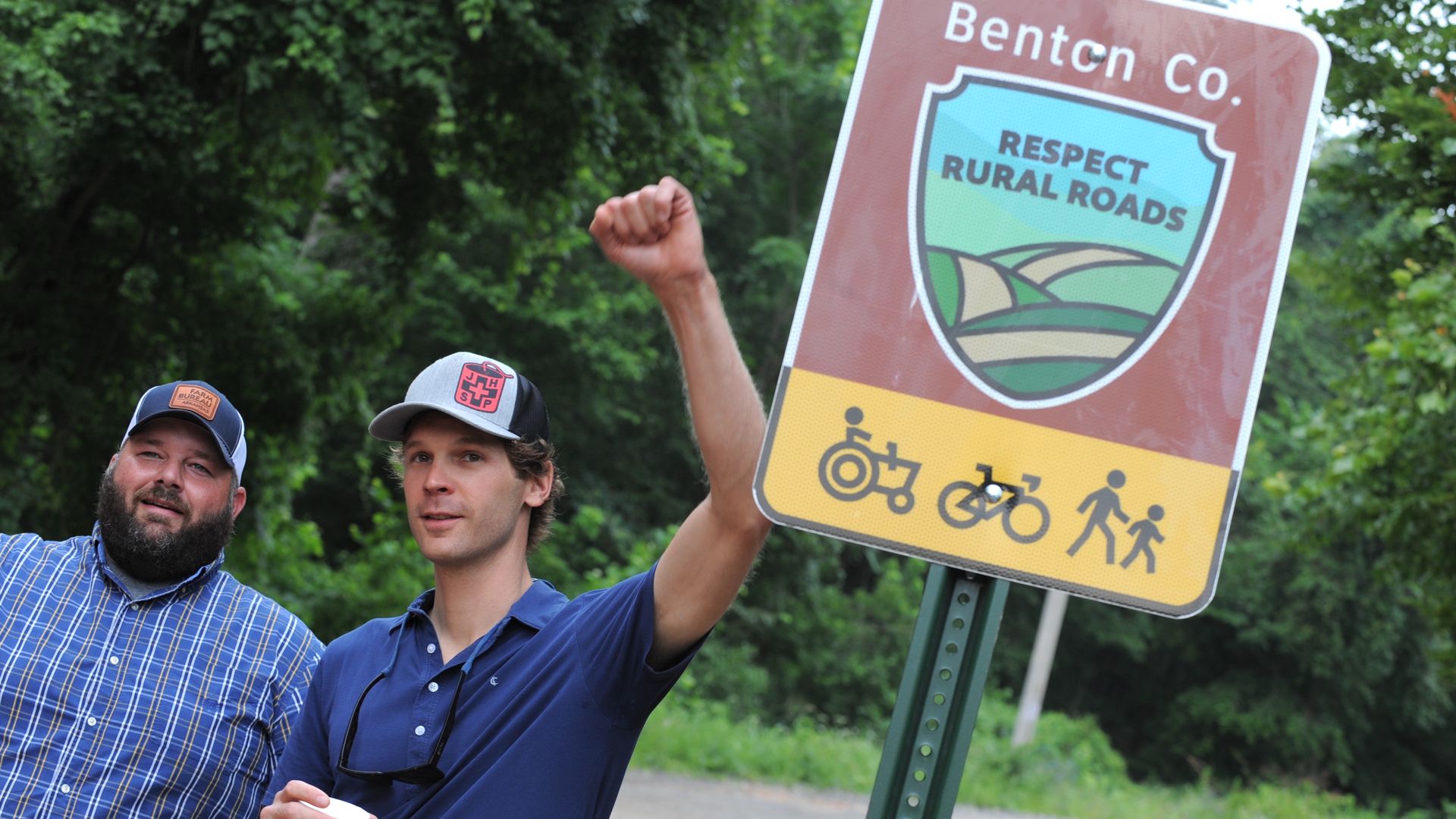 Two men — one with his fist in the air — stand near a sign that reads "Respect Rural Roads."