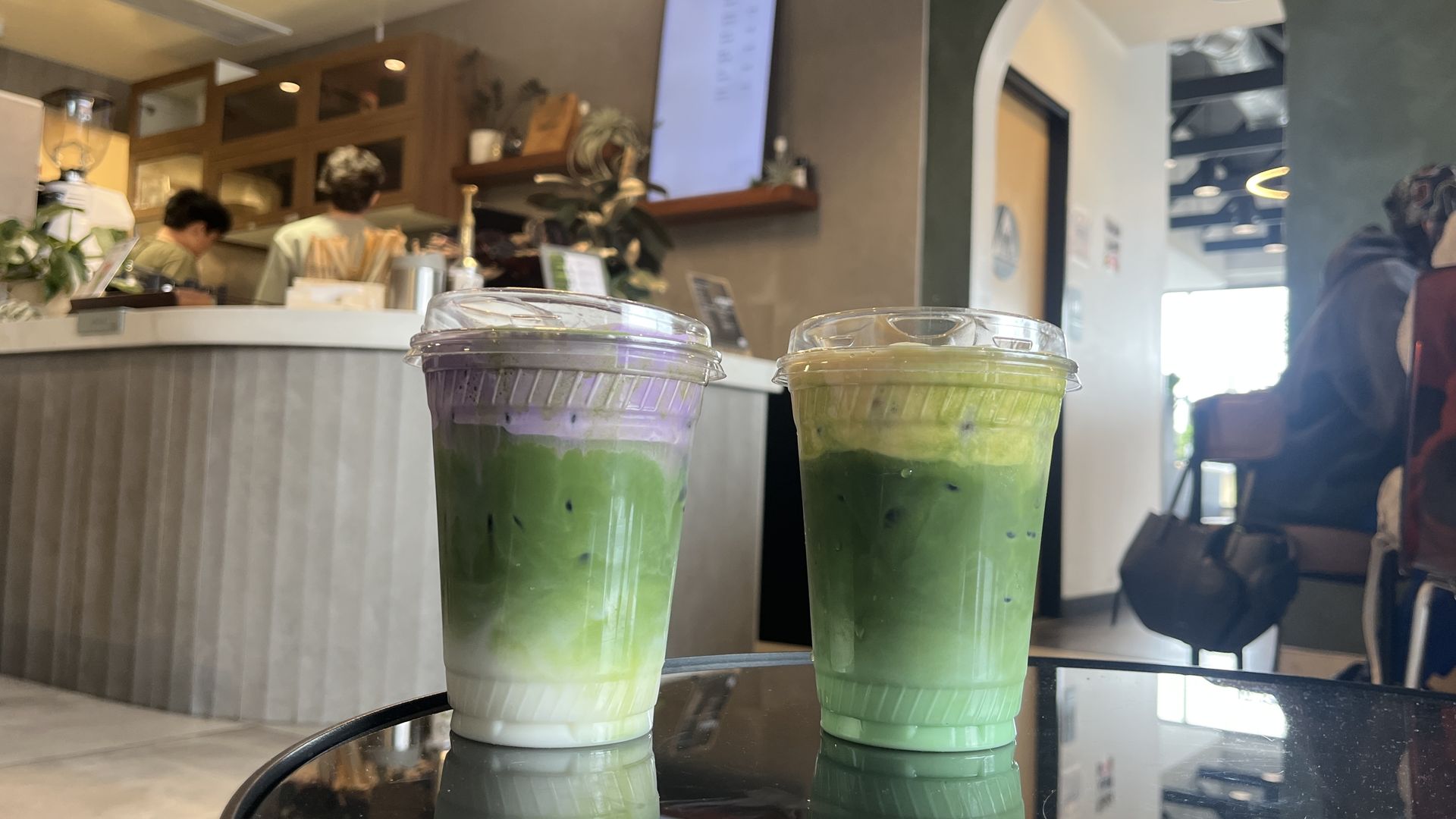 Two iced layered drinks with green, white, and purple/yellow foam tops on a glass table in a cozy cafe with people and shelves in the background.