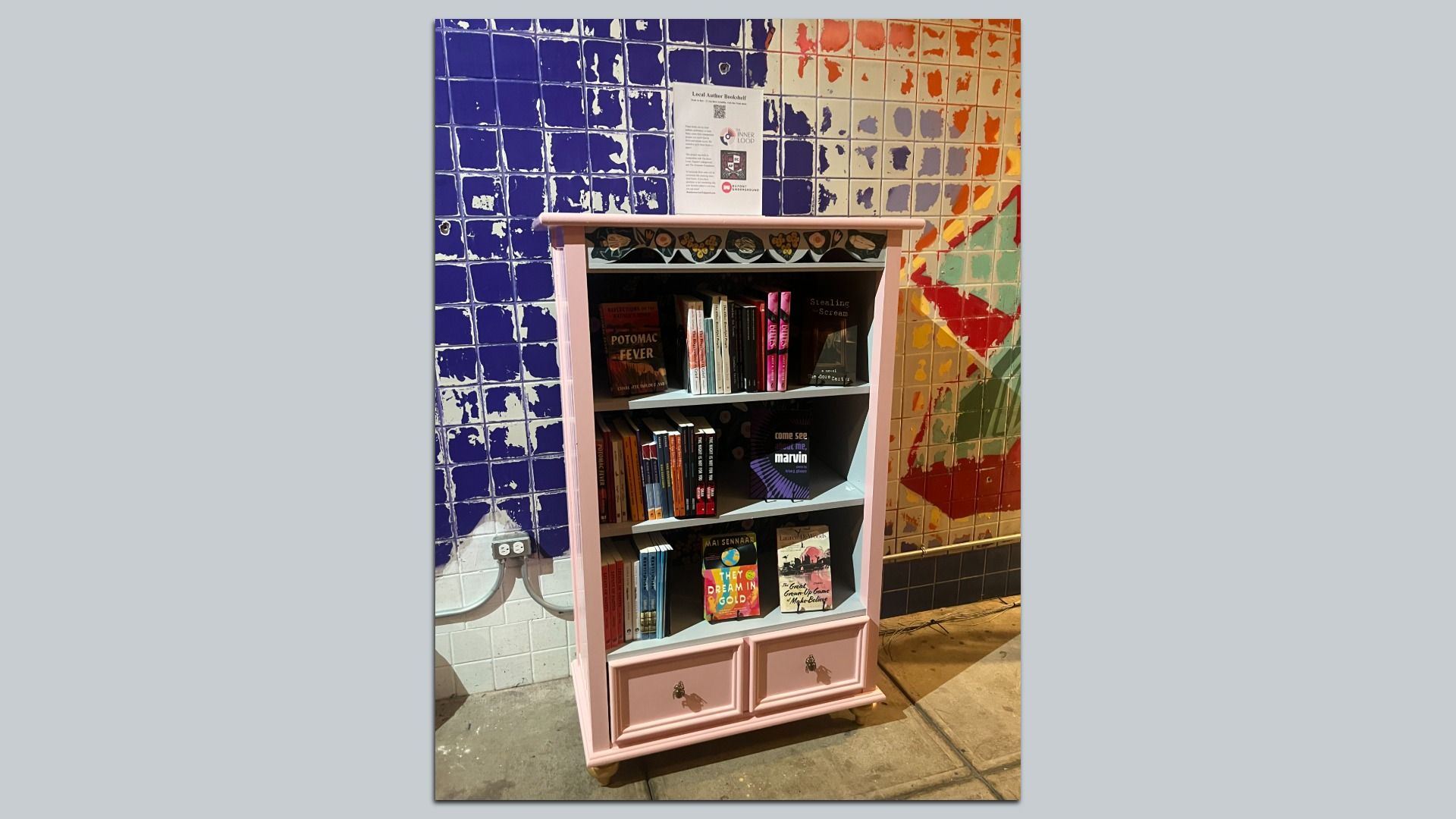A pink vintage bookshelf filled with assorted books stands against a large mosaic tiled wall—left side blue tiles fading to orange and multicolored tiles on the right; a small poster sits above.