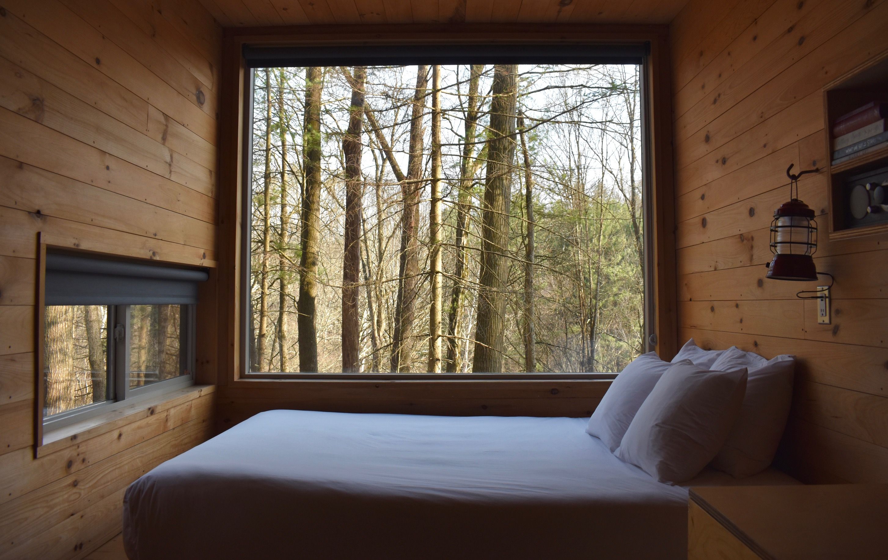 Cozy wooden cabin bedroom with a big window showing a forest. A white bed with pillows sits along the right wall; a small window with a blind is on the left, and a lantern hangs by a shelf.