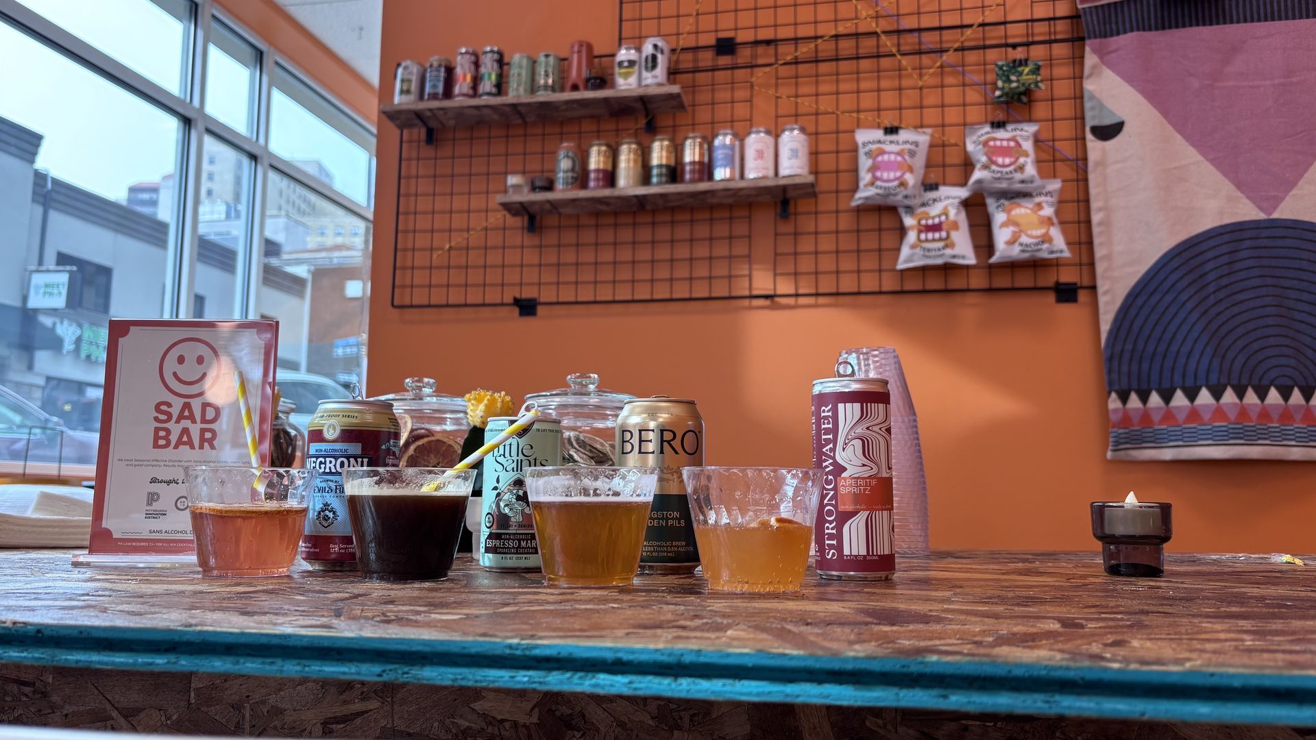 Colorful assorted drinks in glasses and cans on rustic wooden bar with orange wall, snack bags, and window showing city buildings outside.
