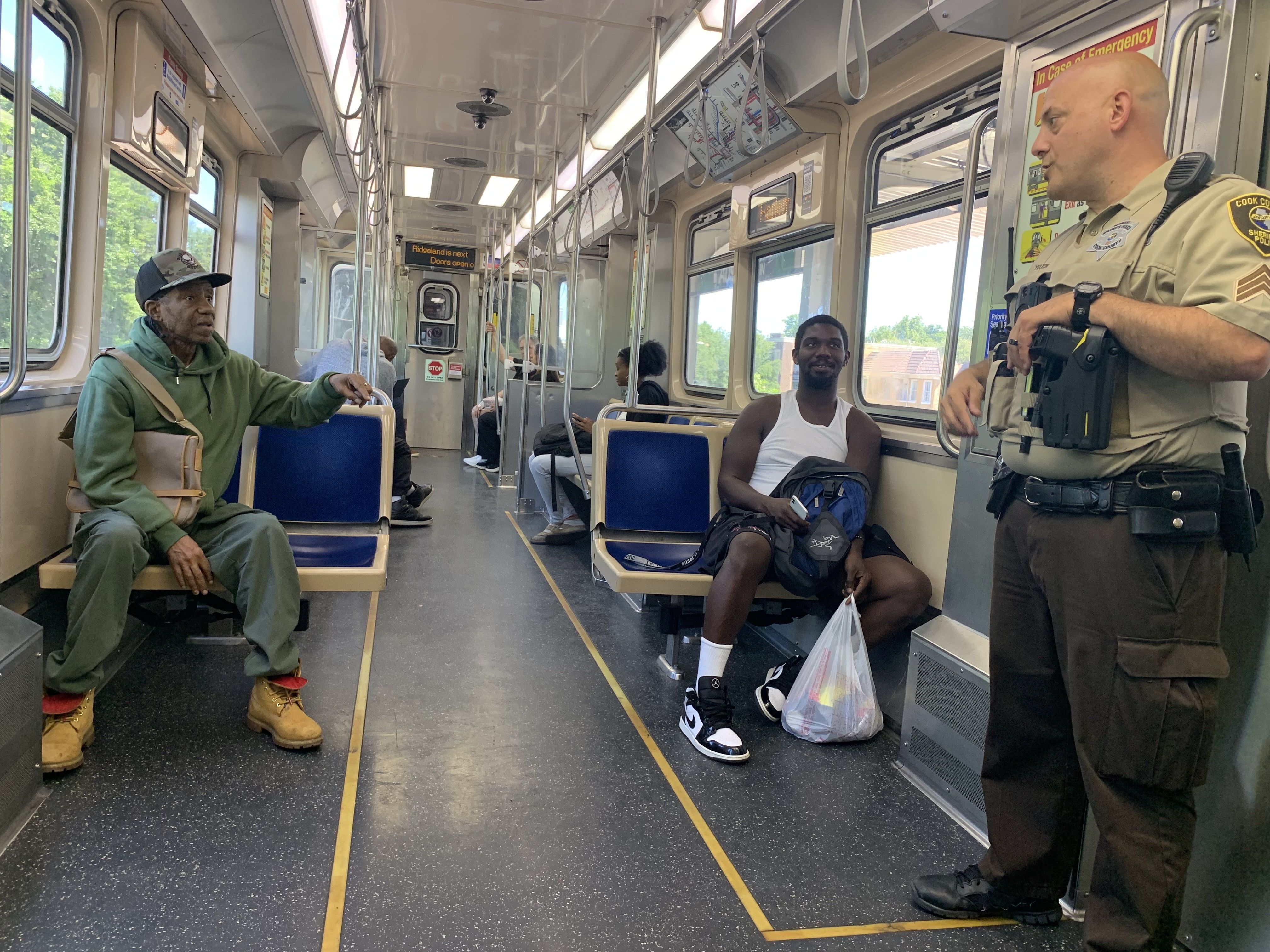 Inside a light subway car with several passengers: left, an older man in a green hoodie and tan boots; center, a man in a white tank top sits; right, a uniformed officer with a radio speaks.