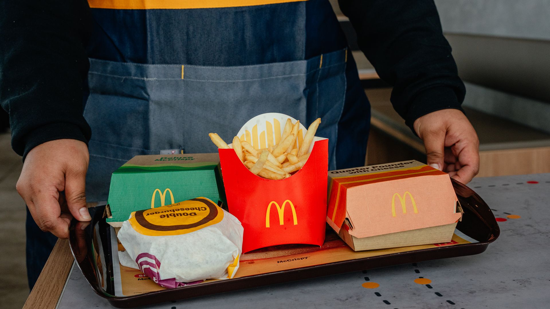 Person wearing a blue denim apron with an orange stripe handles a tray of McDonald's boxes, featuring a red fries container and visible french fries.