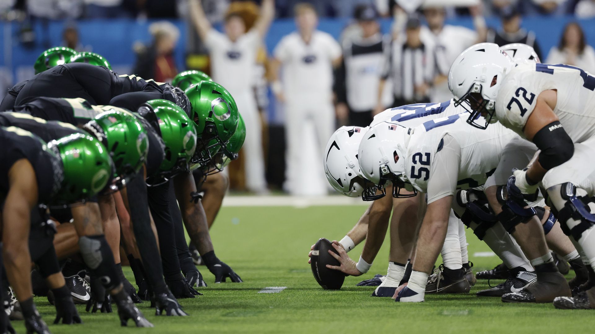 Two American football teams lined up opposite each other at the line of scrimmage; team in black with green helmets and team in white with white helmets, ready to snap the ball.