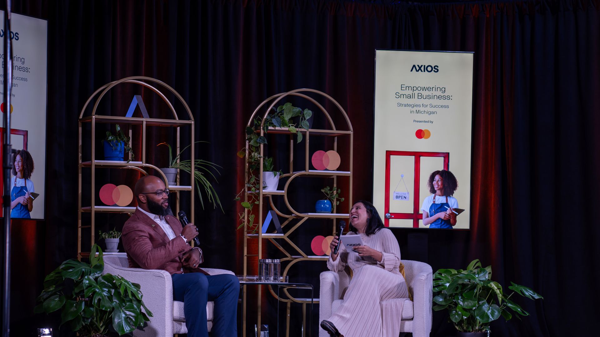 Two people, a man in a brown jacket and a woman in a white dress, sit on stage chairs with microphones, discussing at an Axios event titled "Empowering Small Business: Strategies for Success in Michigan."