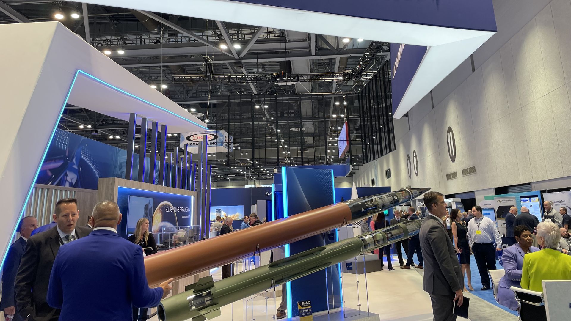 Blue exhibition banner reading LOCKHEED MARTIN with a white star logo hovers above a busy trade show booth; people examine displays while brown-green missiles sit in the foreground.