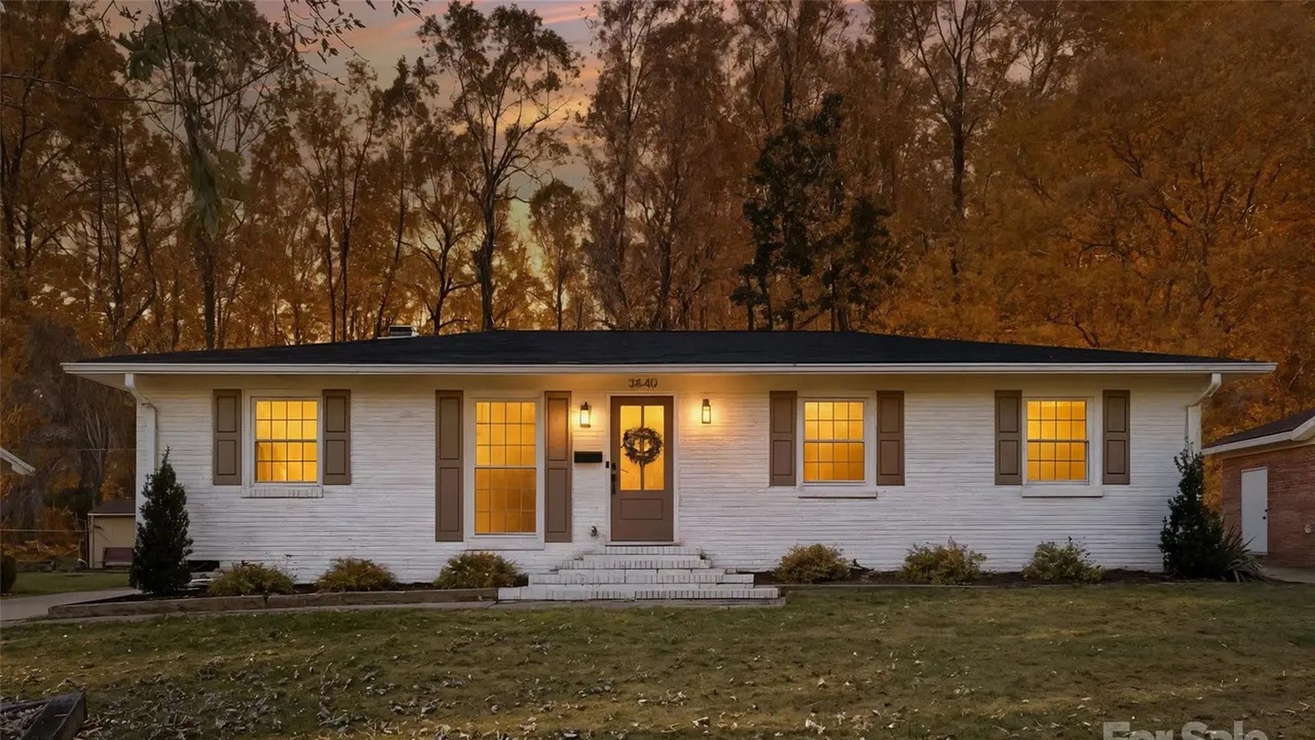 Single-story white brick house with brown shutters and front door wreath, warmly lit windows at dusk, surrounded by trees with autumn foliage, green lawn in front.