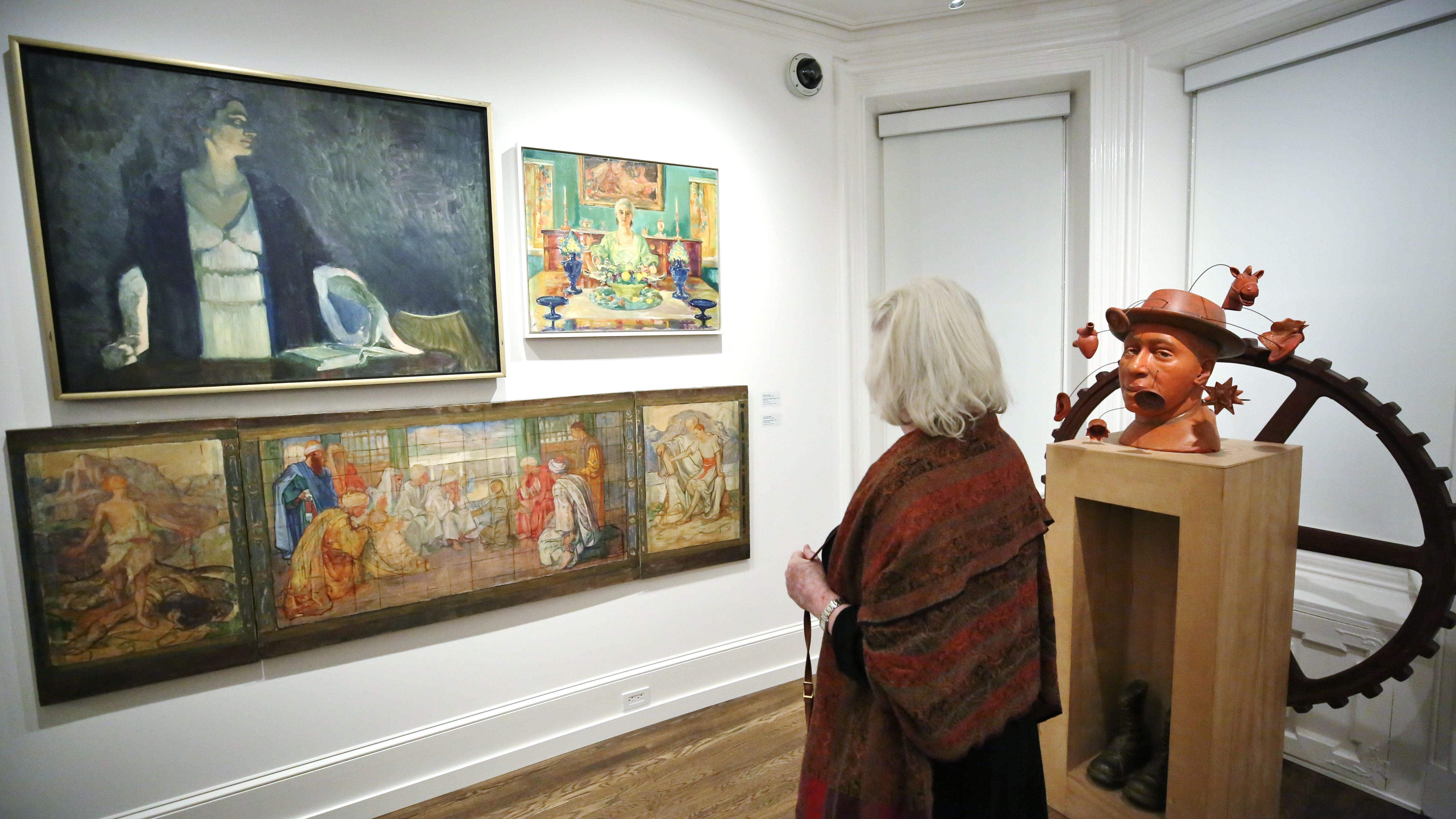 Woman with white hair wearing a brown and red shawl observes paintings and a terracotta sculpture of a man with a hat and boots in an art gallery with white walls and wooden floor.