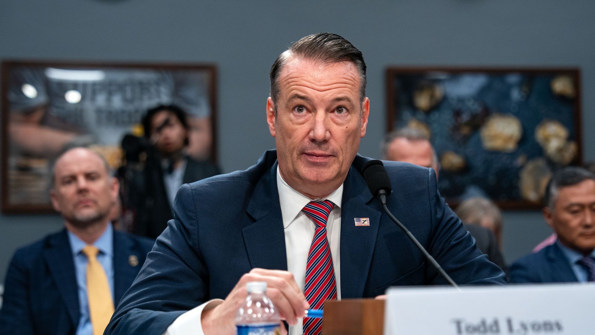 Lyons speaking into a microphone wearing a blue suit jacket with an American flag pen, white collared shirt and red and blue striped tie while holding a pen