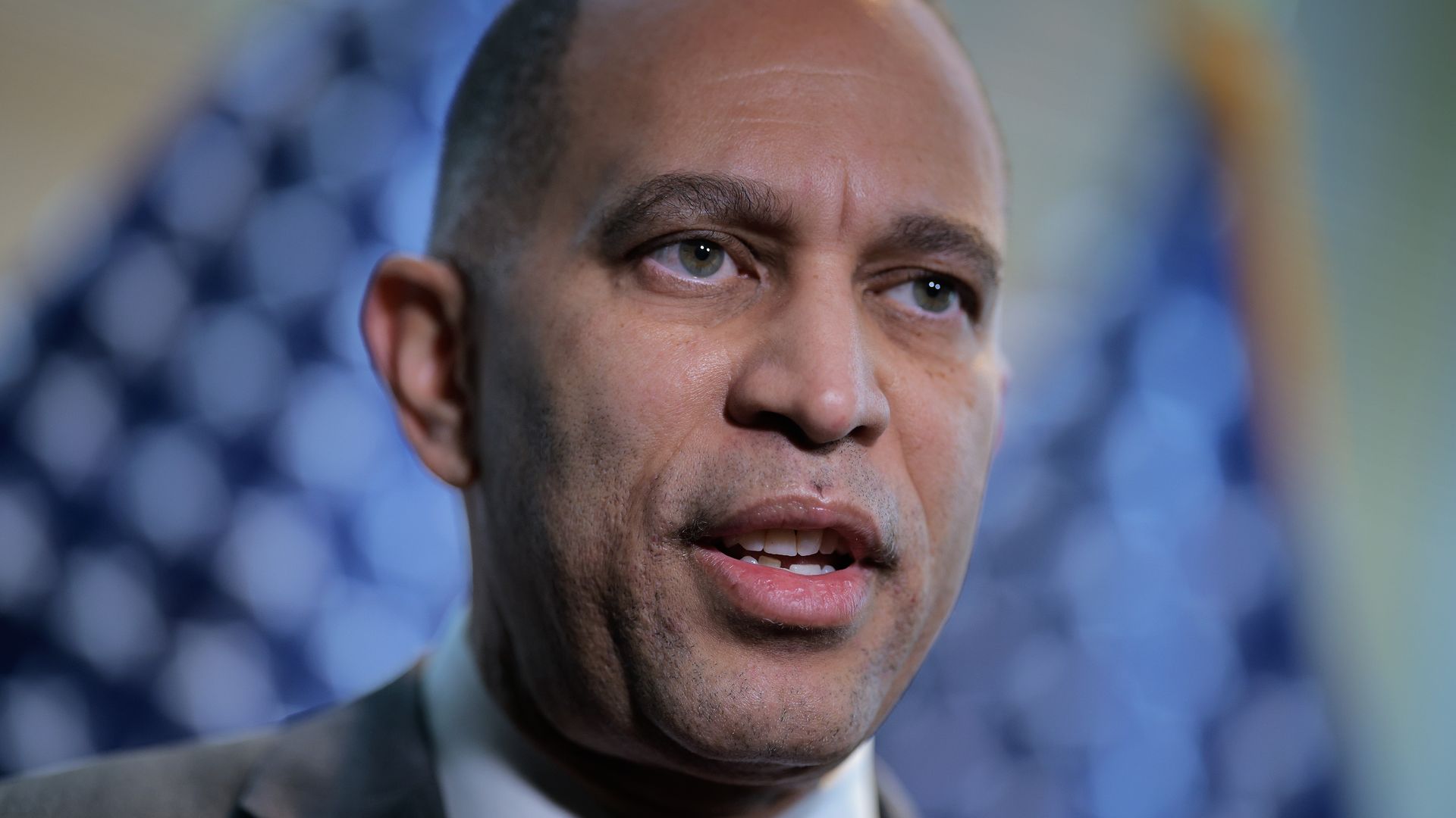 Close-up of a man in a suit and red tie speaking, with a blurred American flag in the background.