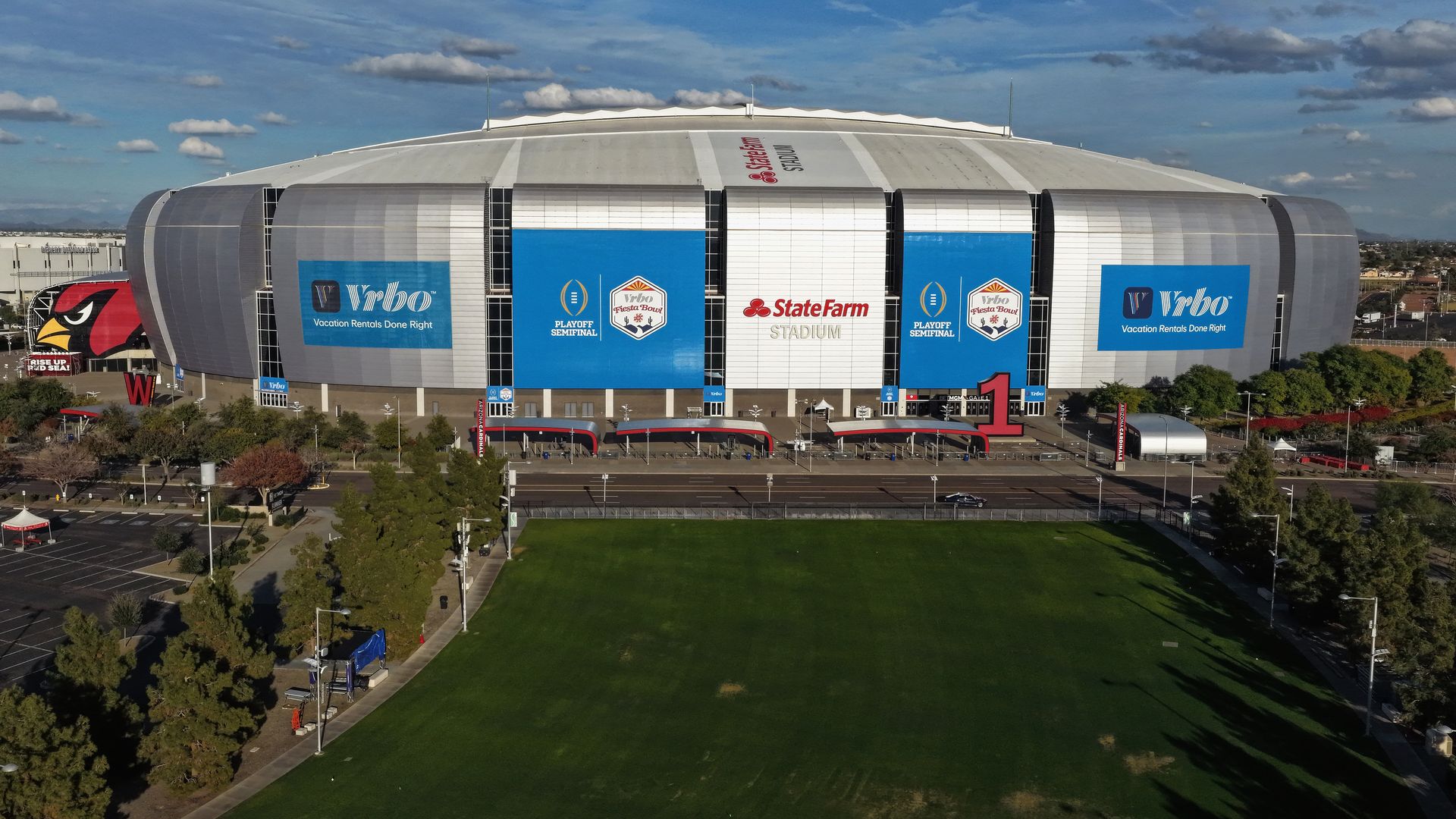 Aerial view of State Farm Stadium with blue and white banners for Fiesta Bowl Playoff Semifinal, surrounded by green grass and trees under a partly cloudy sky.