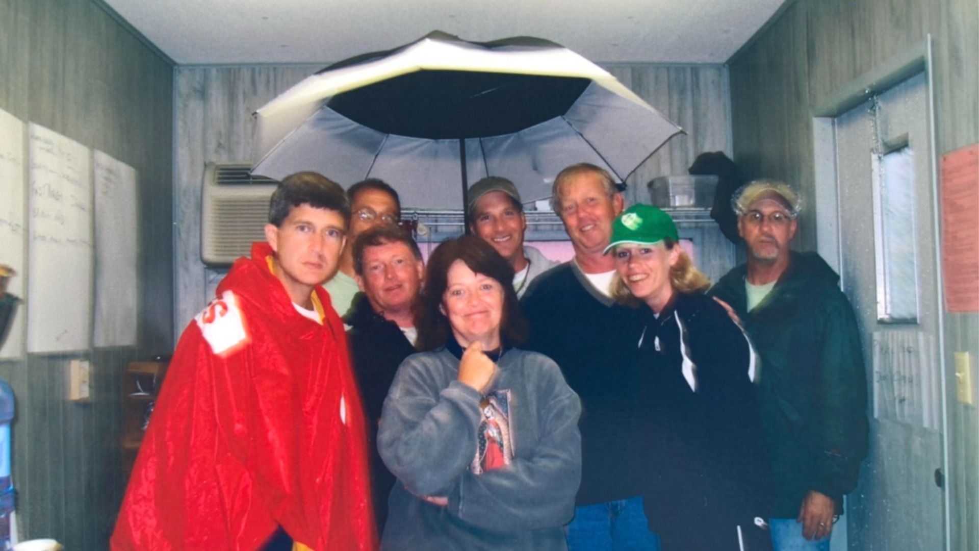 Eight people pose in a small grey metal room. An open umbrella sits overhead. Front center: a woman in a grey sweatshirt; left: a man in a red cape; right: a woman in a green cap; everyone smiles.