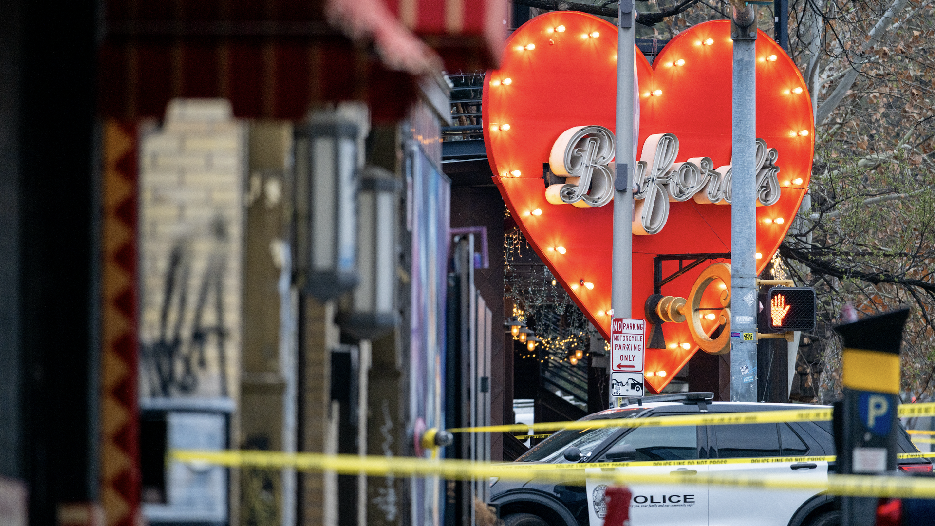 Police car behind yellow tape near a large red heart-shaped sign that reads Biford's, on a city street with fall trees and a traffic signal showing a red hand.