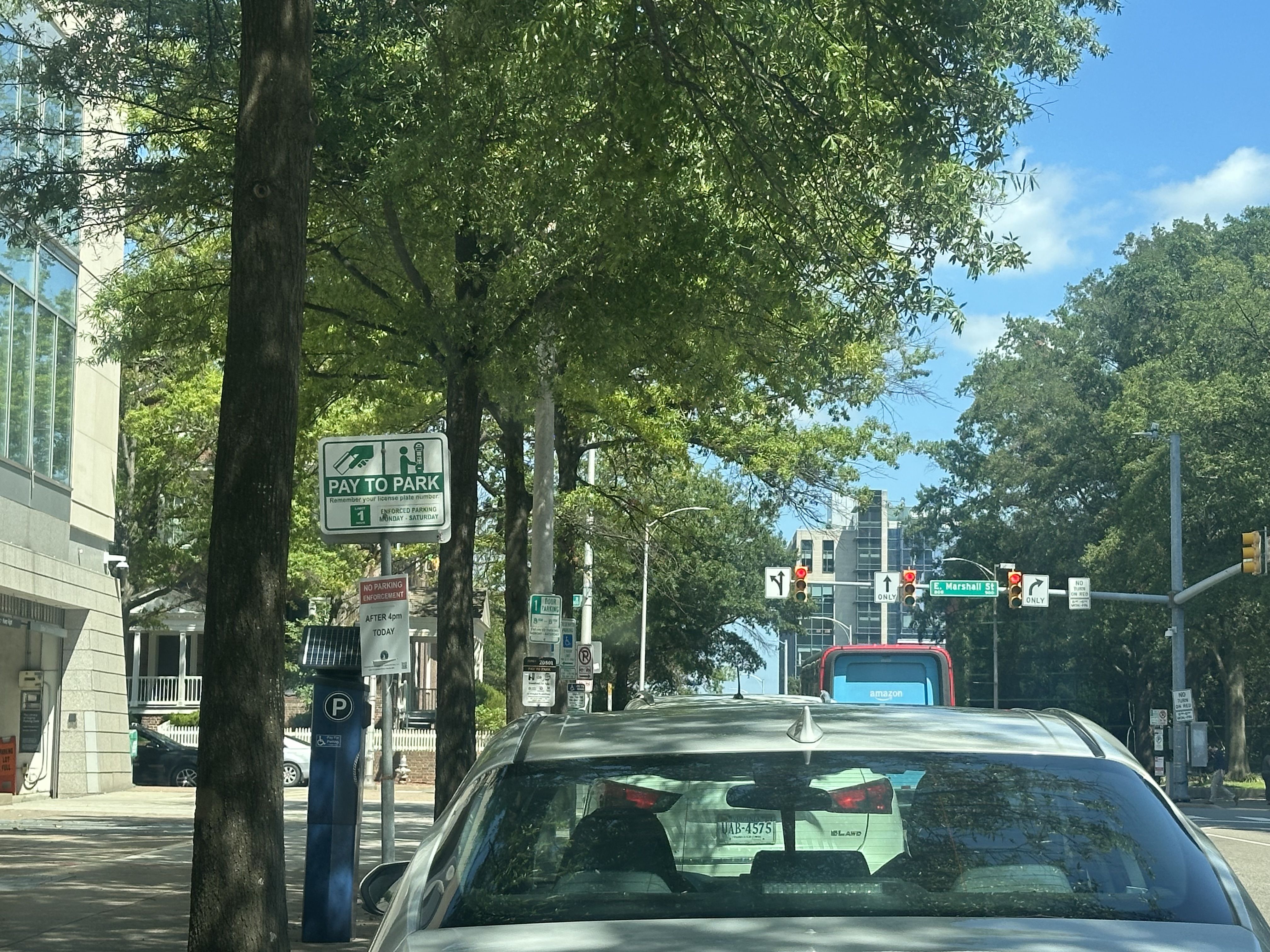 Urban street lined with trees, glass-fronted building on the left, a car in the foreground, and a pay-to-park sign. Traffic lights and street signs ahead under a clear blue sky.