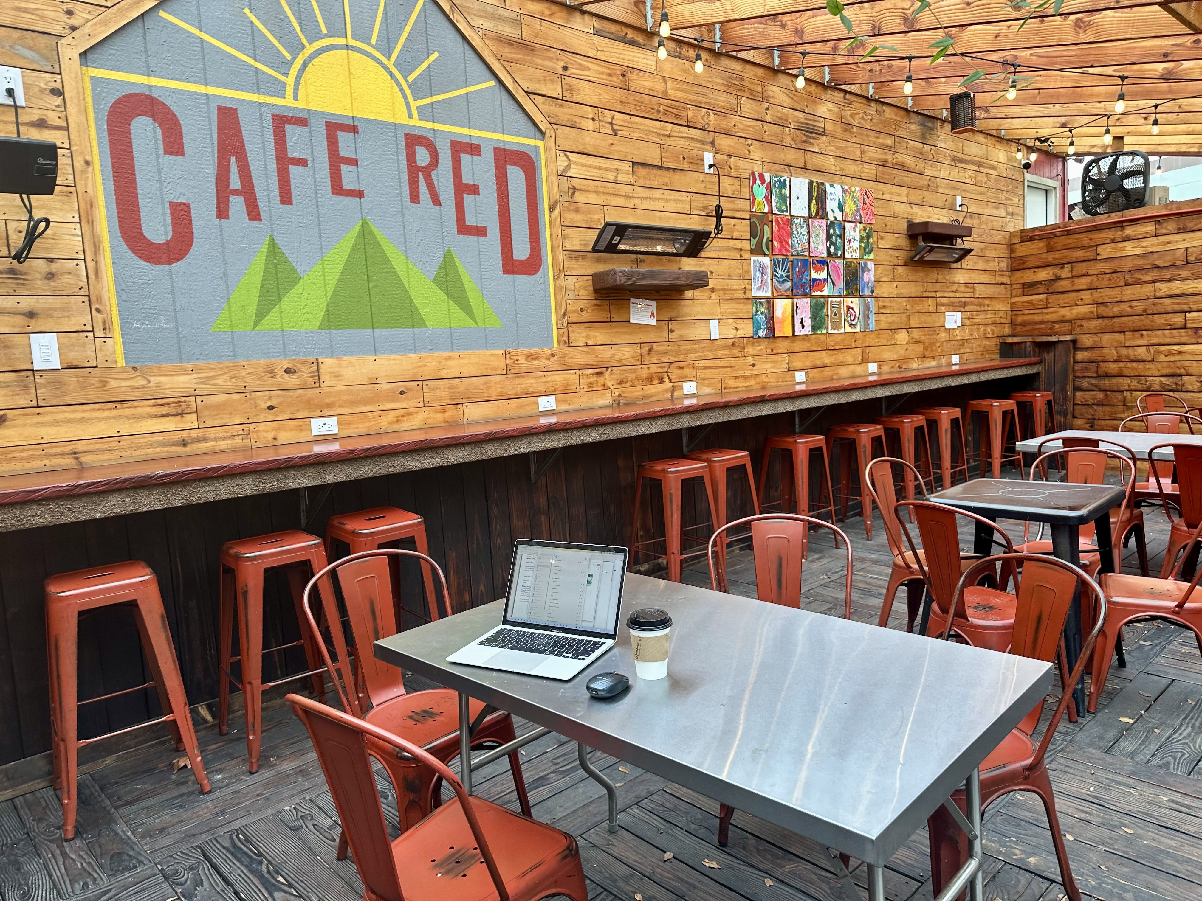 A wood paneled wall with a painting that says "Cafe Red" above barstool seating with outlets, alongside a courtyard with metal tables and red chairs and a laptop sitting on one table.