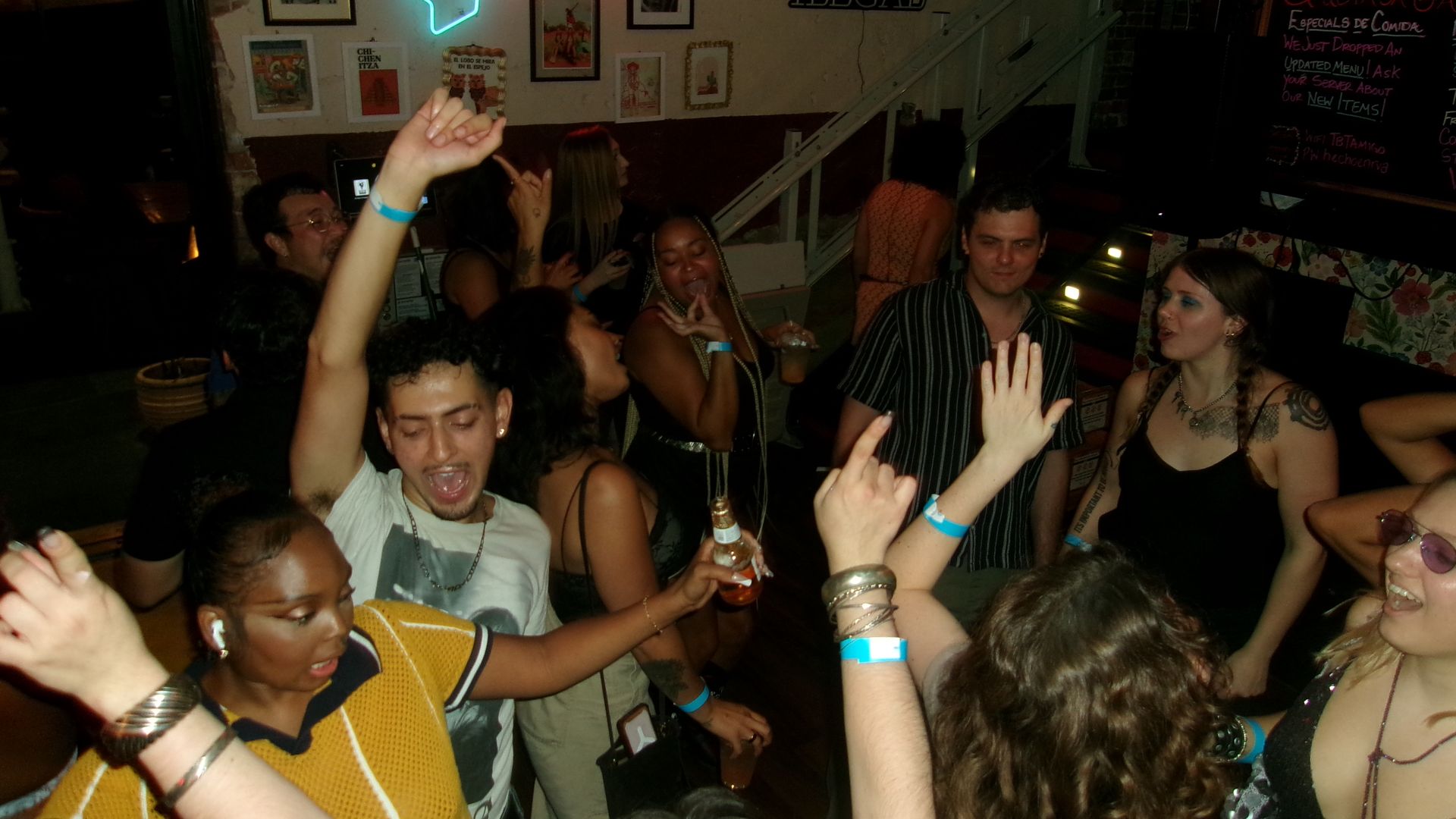 Group of people dancing and enjoying drinks in a dimly lit bar with a neon "Tacos" sign and framed pictures on the wall, creating a lively atmosphere.