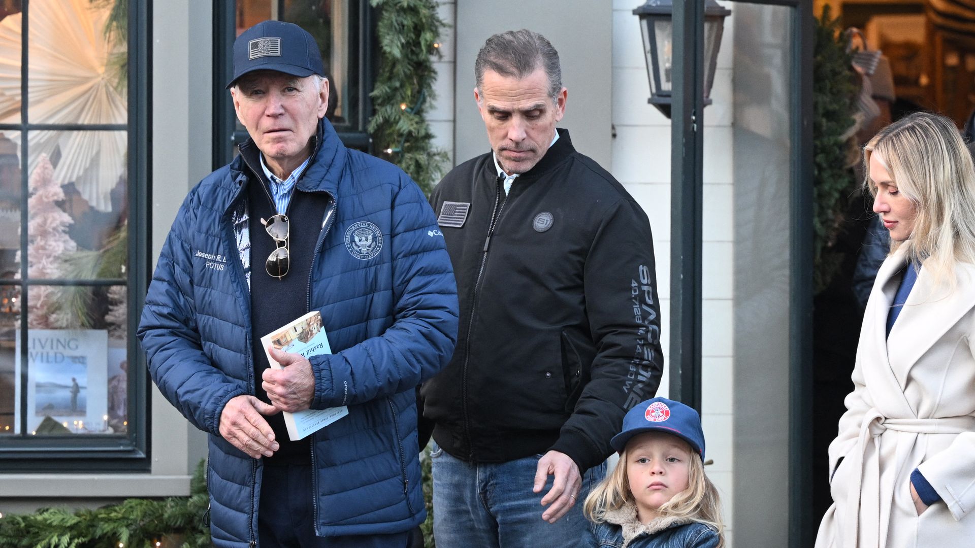 President Biden, wearing a blue puffy jacket, and his son Hunter, wearing a gray windbreaker, with their family outside a white building with black streetlamps.
