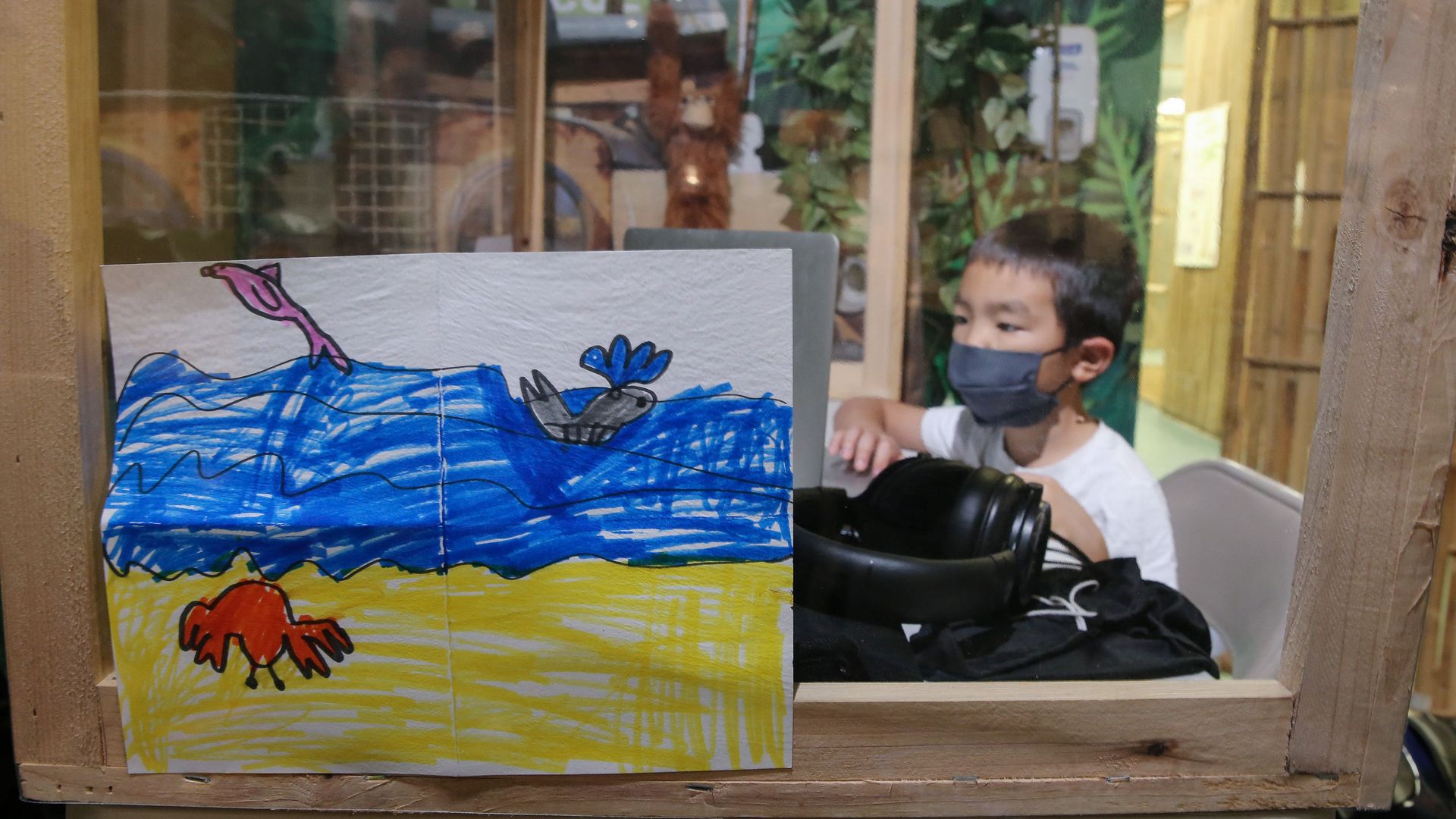 Photo of a drawing with a young student working on their laptop in the background