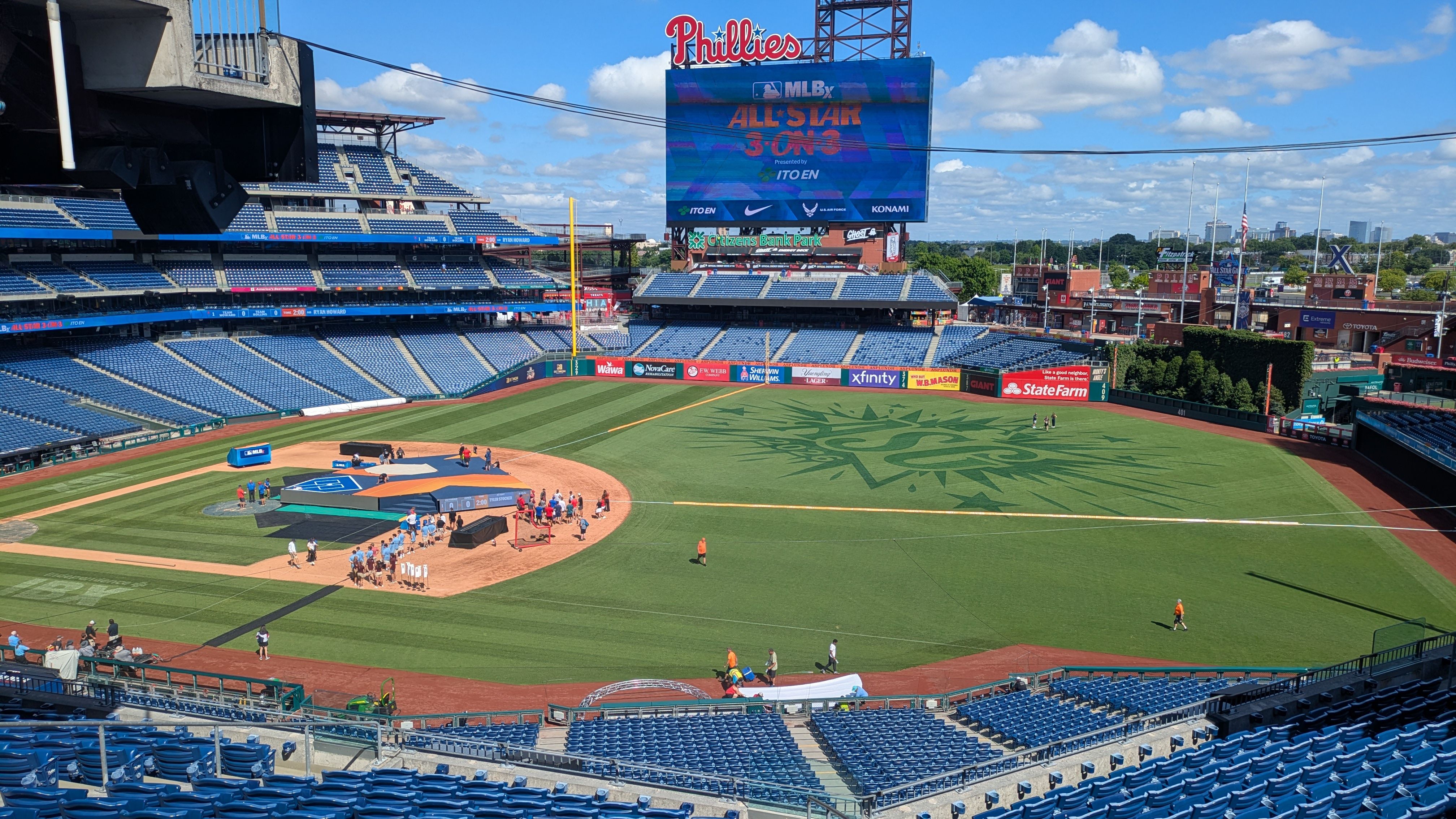 Panoramic view of a Phillies baseball stadium with blue seats, a large video scoreboard displaying ALL-STAR 3 ON 3, a bright blue sky, and players on the infield.